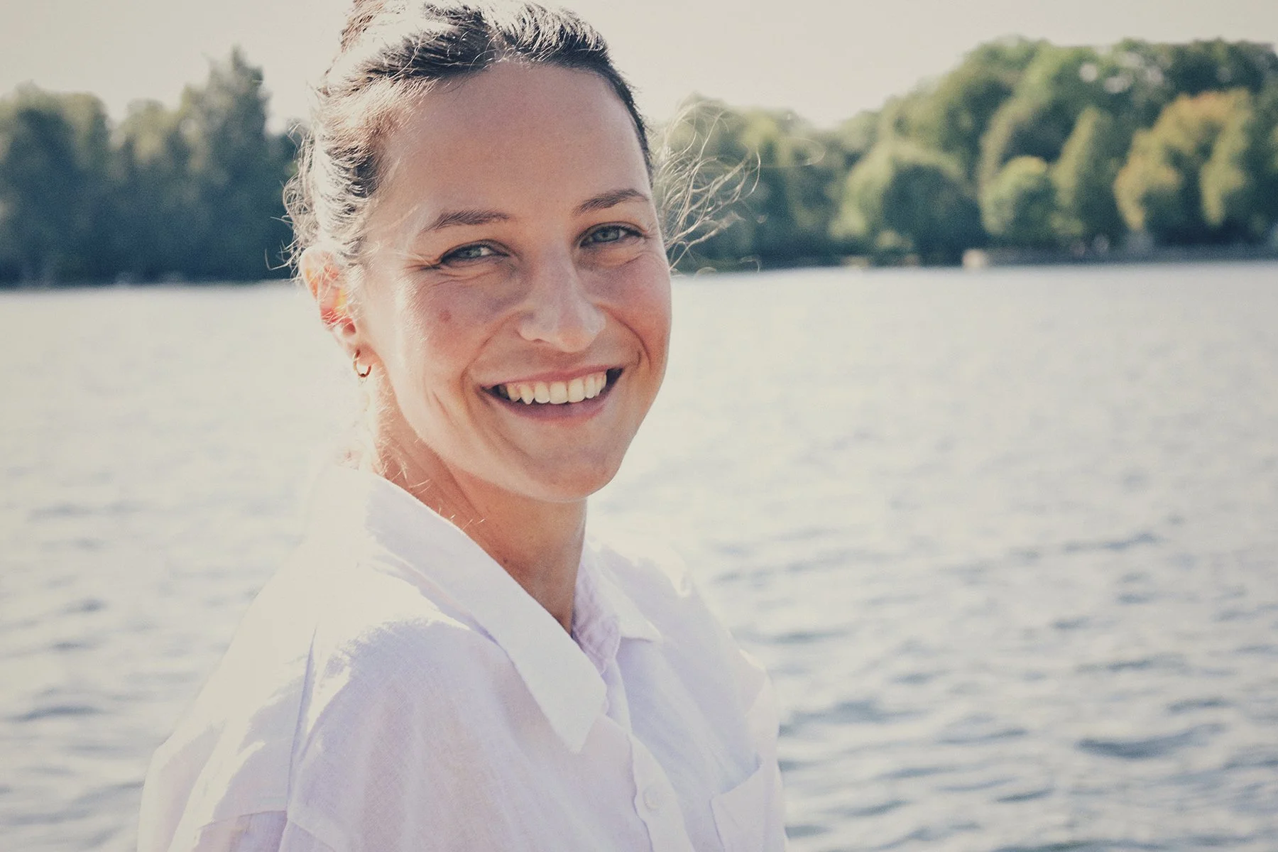 Lächelnde junge Frau mit braunen Haaren und blauen Augen, trägt weiße Kleidung, sitzt am Wasser mit grünen Bäumen im Hintergrund, sonniger Tag.