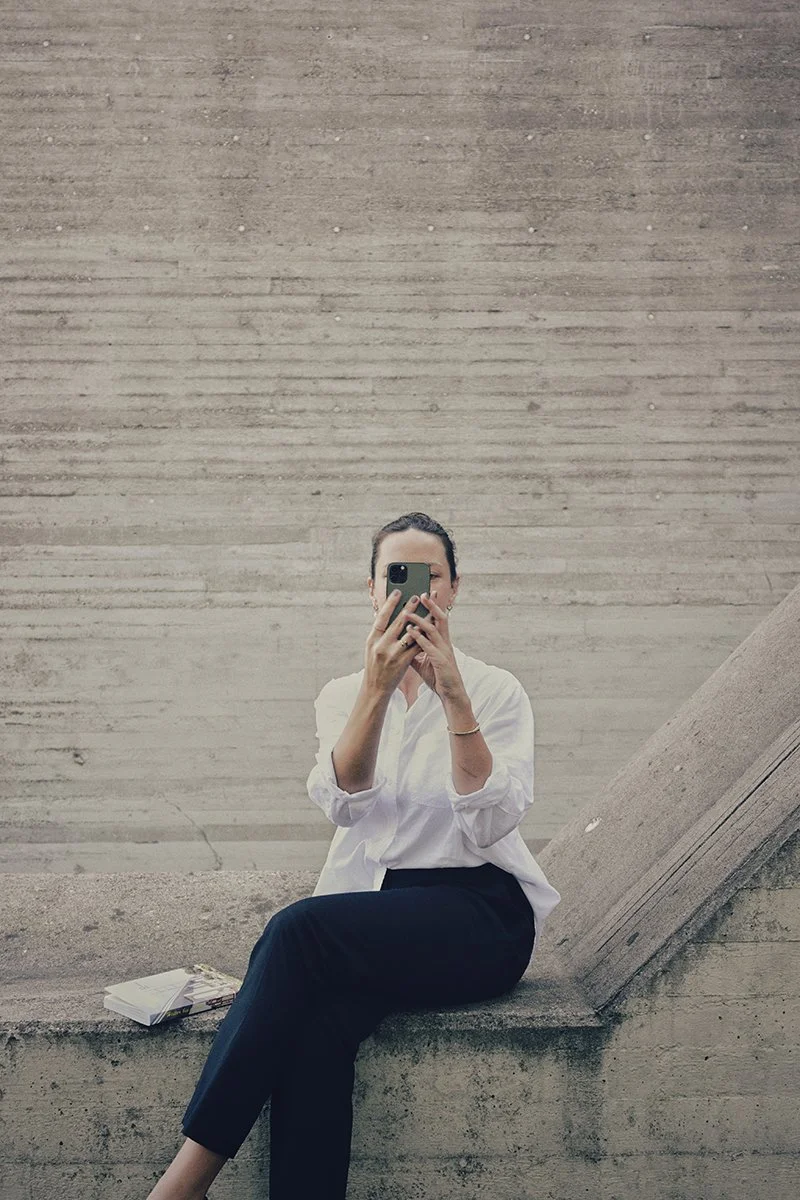 Eine Frau sitzt auf einer Betonbank vor einer modernen, grauen Betonmauer und nimmt ein Selfie mit einem Smartphone auf.