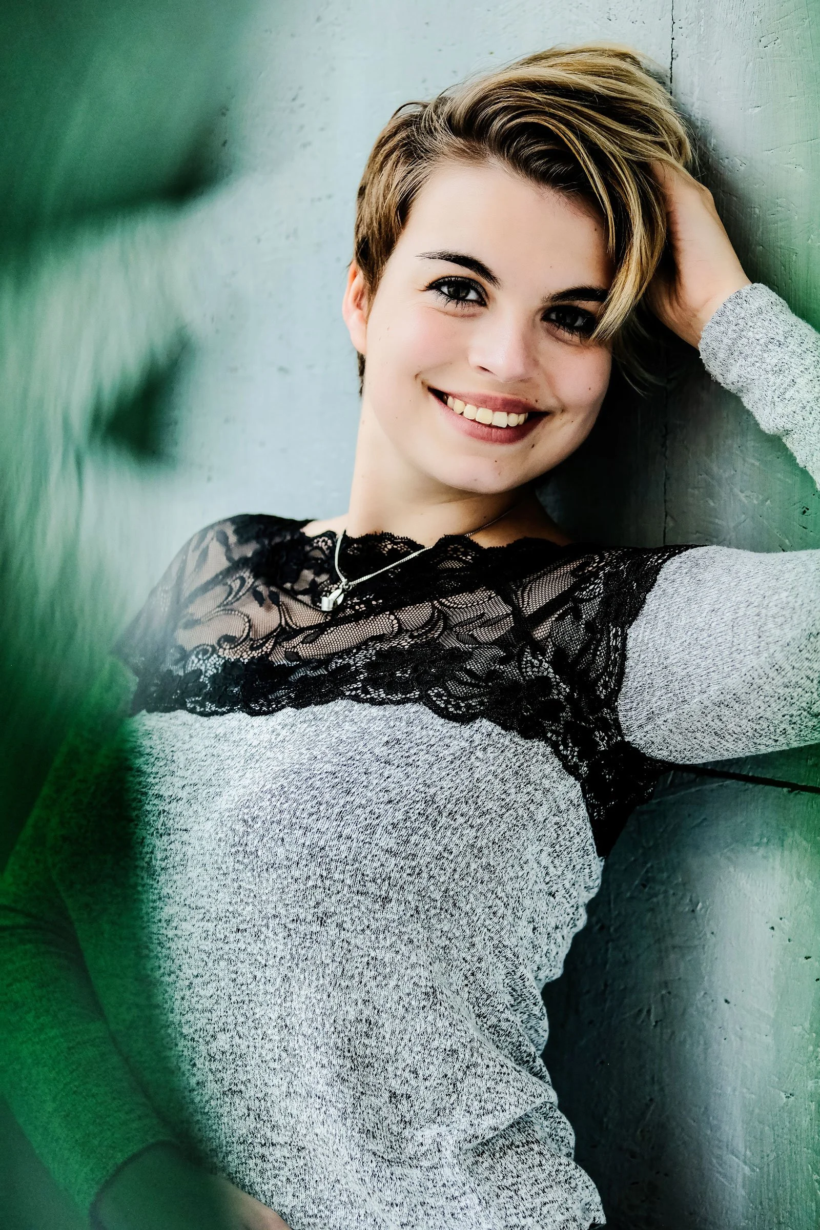 Junge Frau mit kurzen, gestylten Haaren, grauem Pullover mit schwarzen Lace-Akzenten, lächelnd und die Hand im Haar, vor grauem Betonwand sitzend.
