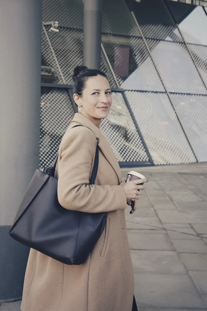 Eine Frau in einem beige Mantel, die eine Tasse Kaffee hält, vor einem modernen Gebäude.