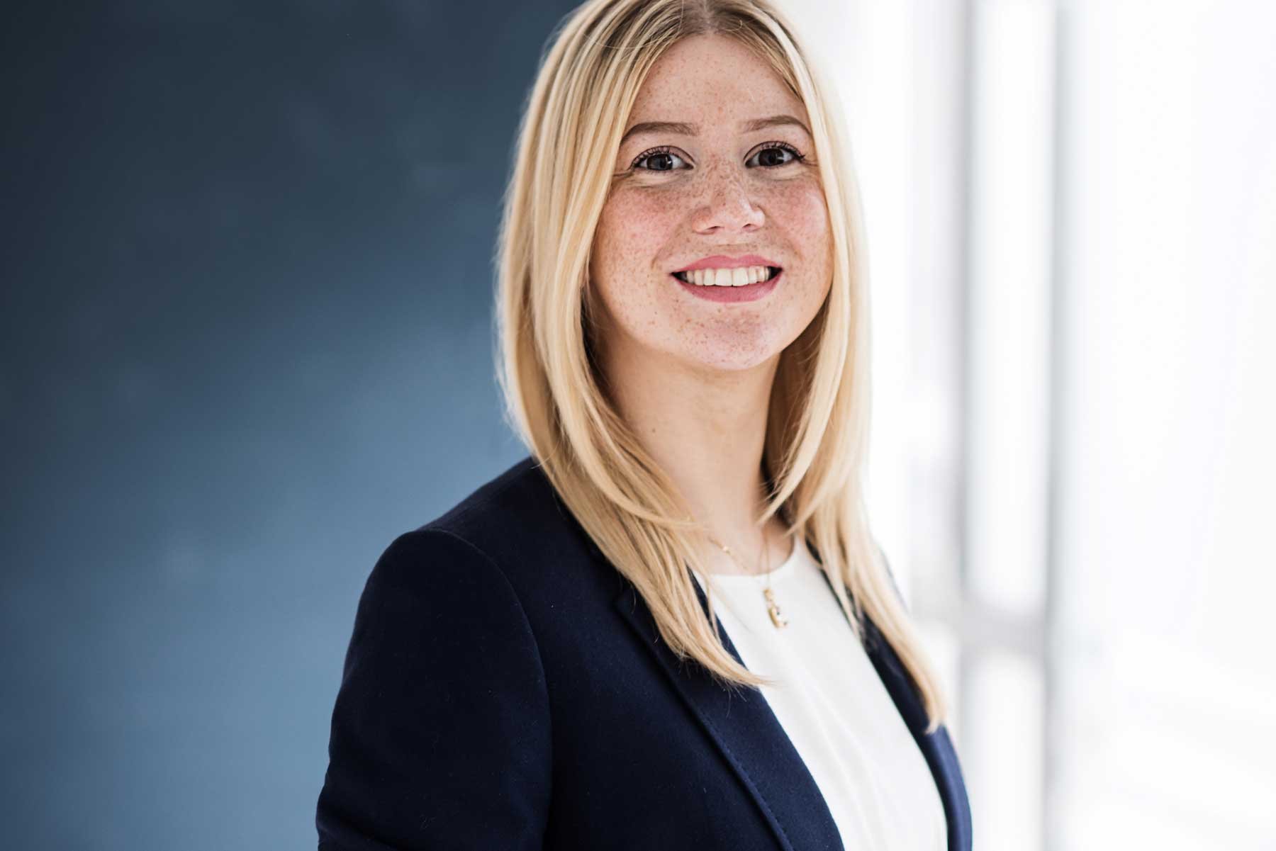 Das Bild zeigt eine blonde Frau in Geschäftskleidung, die vor einem modernen, hell erleuchteten Hintergrund steht und in die Kamera lächelt. Es ist ein professionelles Bewerbungsfotos im Fotostudio in Hannover