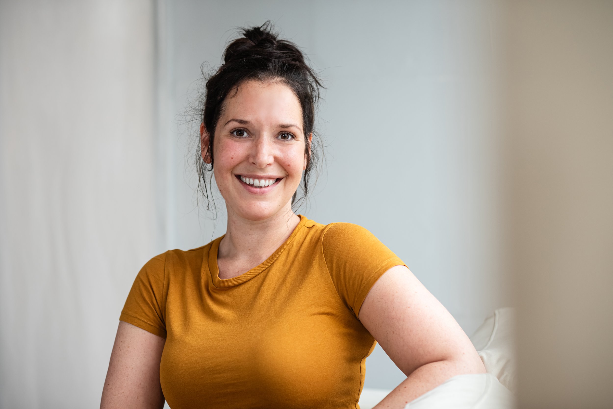 Eine lachende junge Frau mit dunklem Haar in einem Haarknoten, trägt ein orangefarbenes T-Shirt, sitzt auf einem weißen Sofa vor einem unauffälligen grauen Hintergrund. Professionelle Bewerbungsfotos im Fotostudio in Hannover