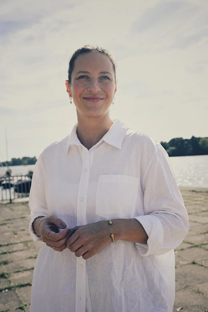 Eine Frau trägt ein weißes Hemd und steht am Wasser mit einem sonnigen Himmel im Hintergrund.