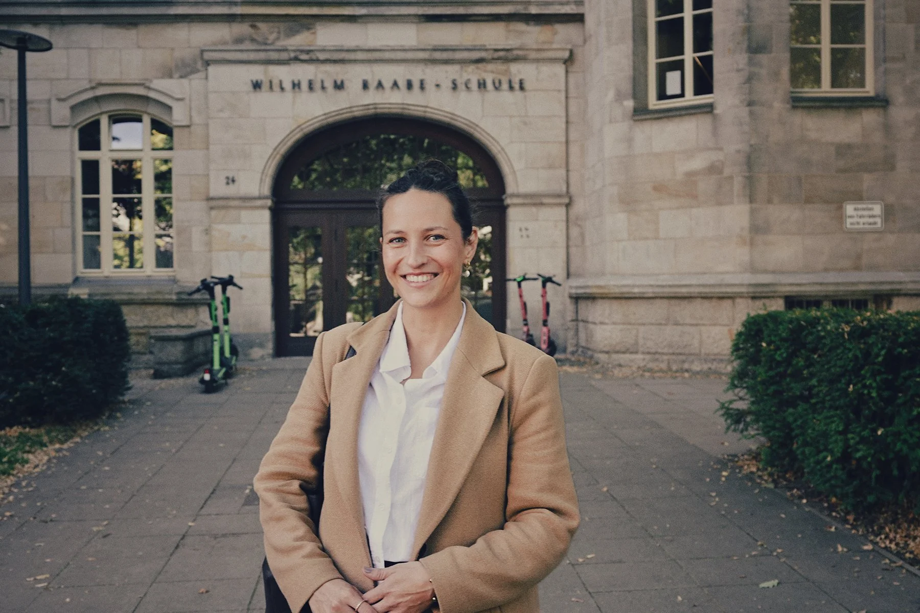 Eine Frau steht vor der Wilhelm Raabe Schule, trägt einen beige Mantel und eine weiße Bluse, im Hintergrund sind Scooter und Bäume sichtbar.