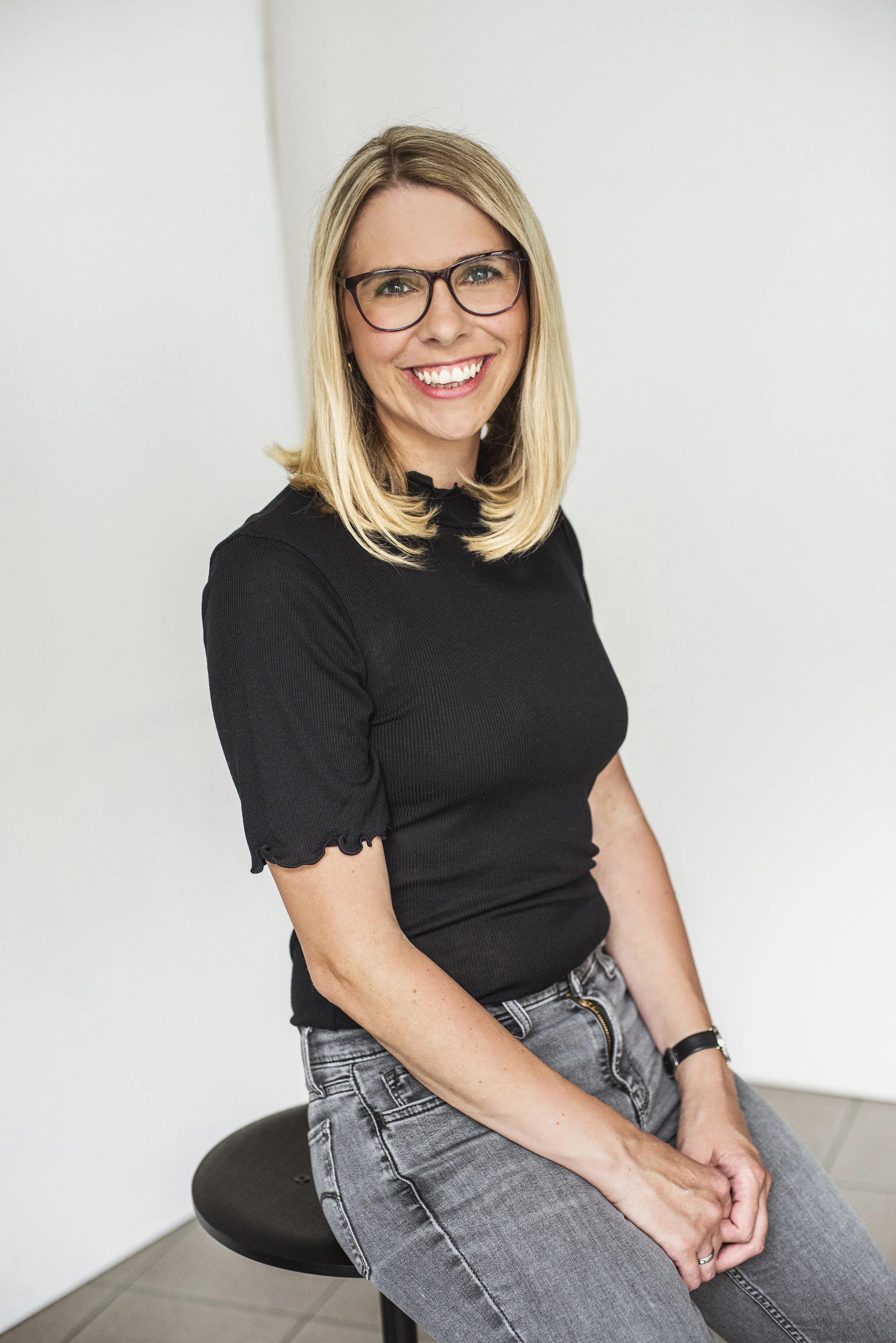 Professionelles Bewerbungsfotos im Fotostudio in Hannover. Eine junge Frau mit Brille, lächelnd, sitzt auf einem schwarzen Hocker vor hellen Wand. Das Bild sieht sehr natürlich aus