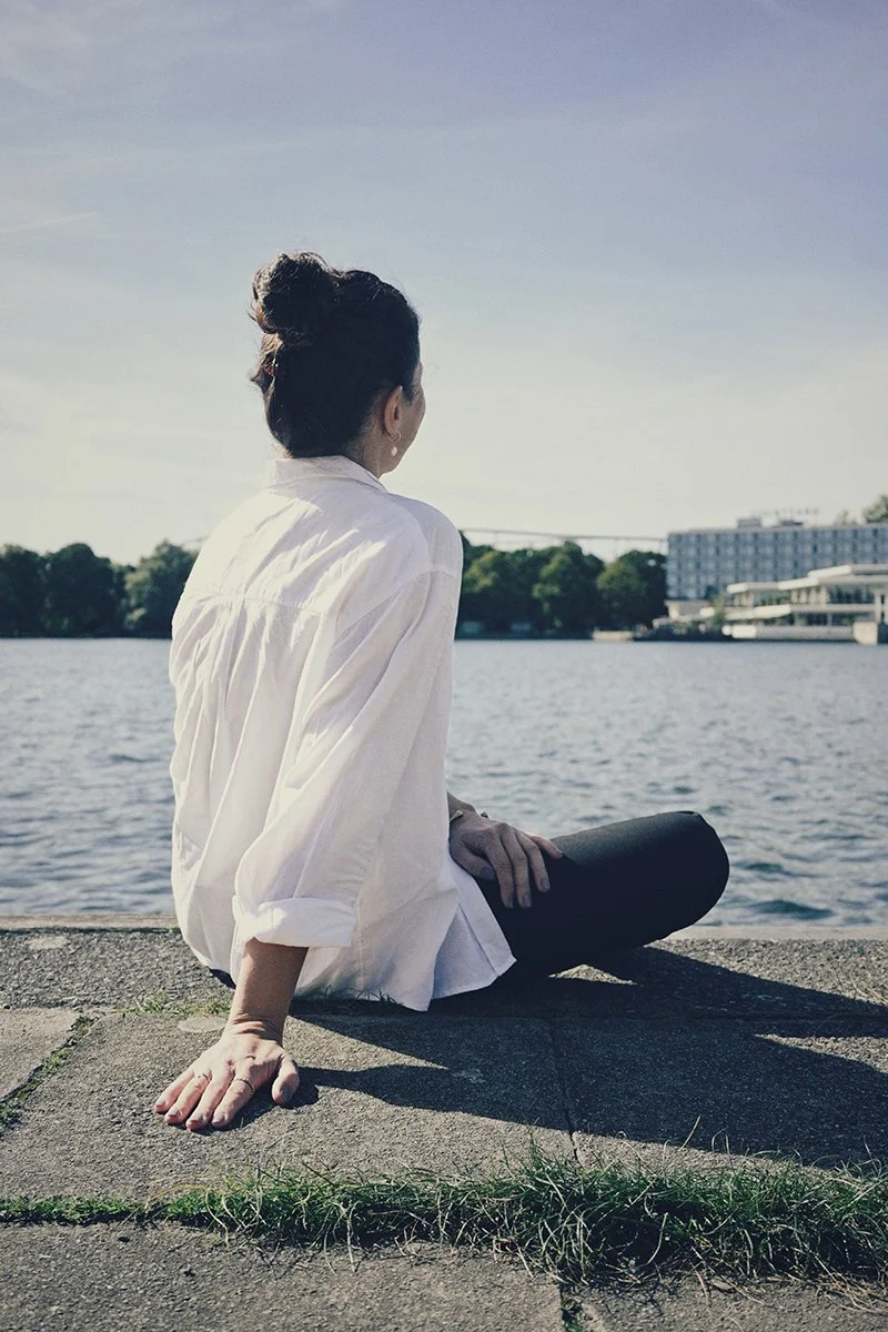 Frau sitzt am Ufer eines Flusses oder Sees, Blick ins Wasser gerichtet, während die Sonne scheint, mit urbaner Hintergrundkulisse.