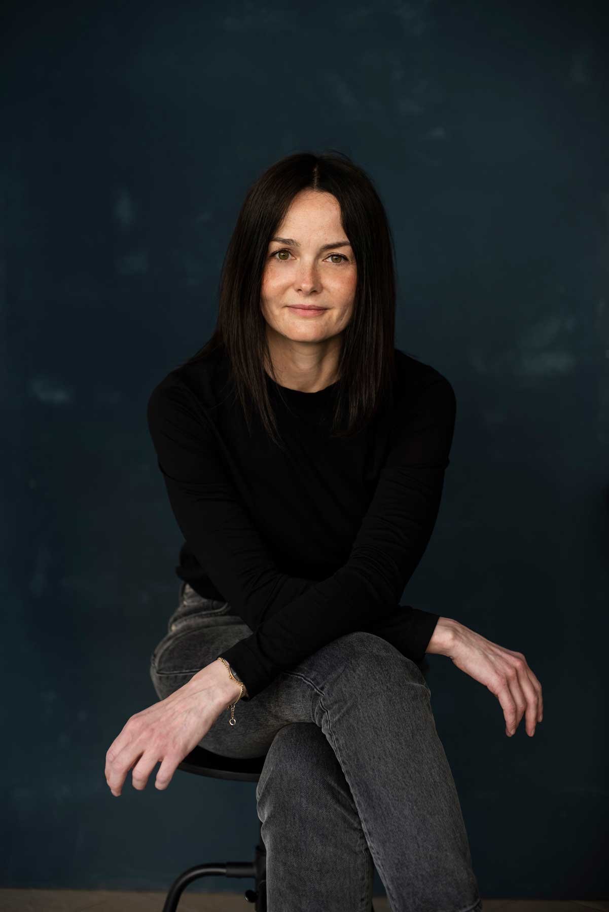 Die selbe Frau wie mit der grünen Jacke und schwarzen Haaren sitz vor dunklem Hintergrund, trägt schwarzes Oberteil und graue Jeans. Professionelle Bewerbungsfotos im Fotostudio in Hannover das sehr cool aussieht