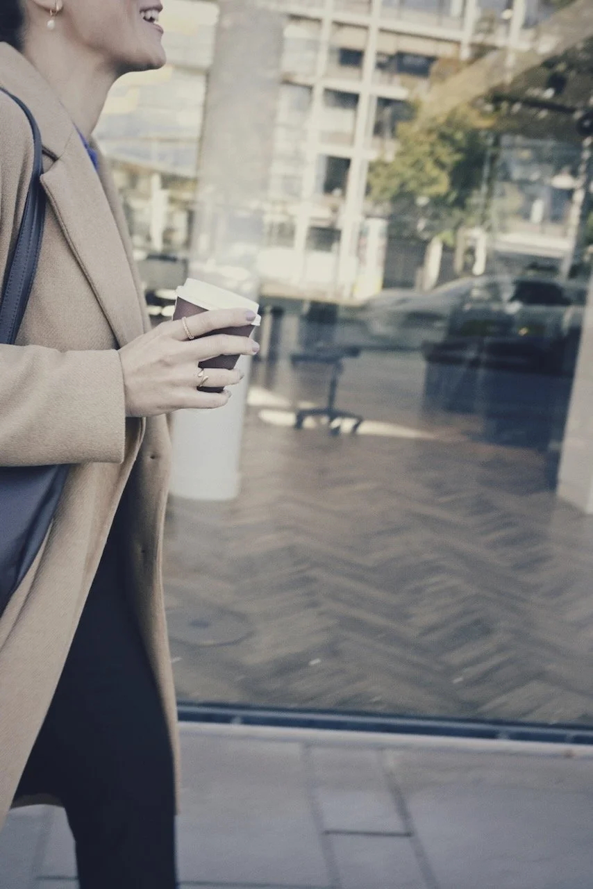 Eine Frau hält einen Pappbecher mit Getränk, trägt einen beige Mantel und eine Umhängetasche, steht vor einer reflektierenden Glasscheibe, im Hintergrund sind Stadtgebäude und Bäume sichtbar.