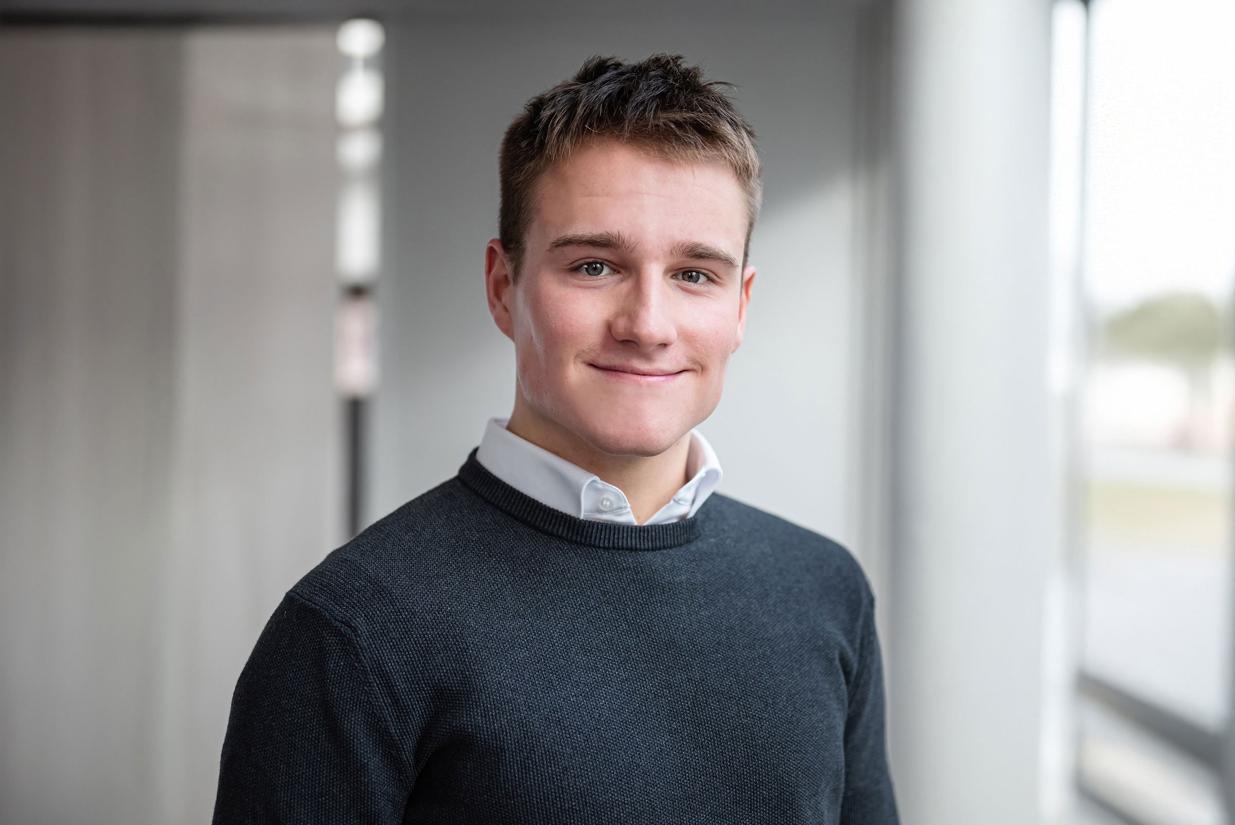 Ein junger Mann mit kurzen braunen Haaren, einem weißen Hemd und einem dunkelgrauen Pullover steht in einem hell erleuchteten Raum mit großen Fenstern. Professionelle Bewerbungsfotos im Fotostudio in Hannover