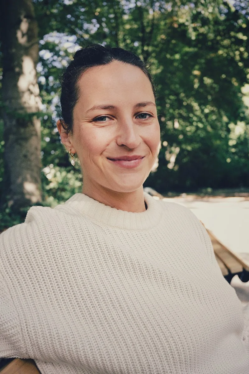 Nahaufnahme einer lächelnden Frau mit dunklem Haar, die im Freien sitzt, umgeben von Bäumen.