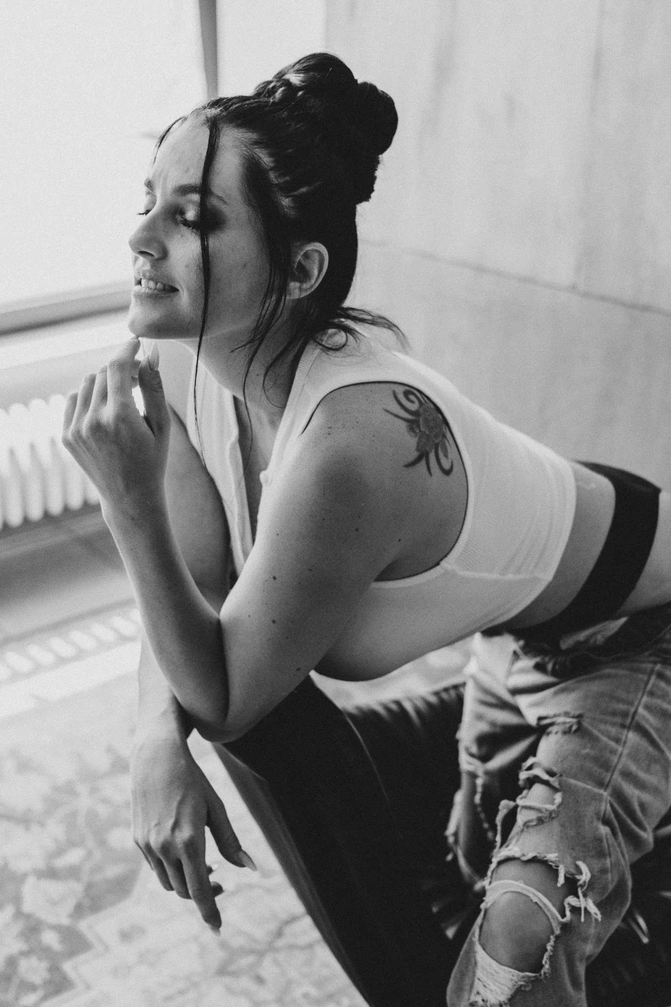 Schwarz-weiß-Foto einer lächelnden Frau mit dunklen Haaren in einem Dutt, die in einem Raum sitzt. Sie trägt ein Tank-Top und vollzogene zerrissene Jeans. Sie hat eine Tätowierung auf ihrem Schulterarm.