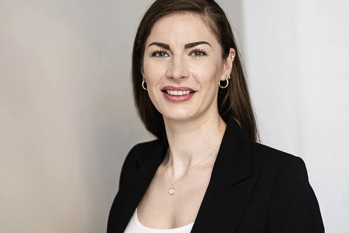 Porträt einer lächelnden Frau mit dunklem Haar in einem schwarzen Blazer vor einem weißen Hintergrund.