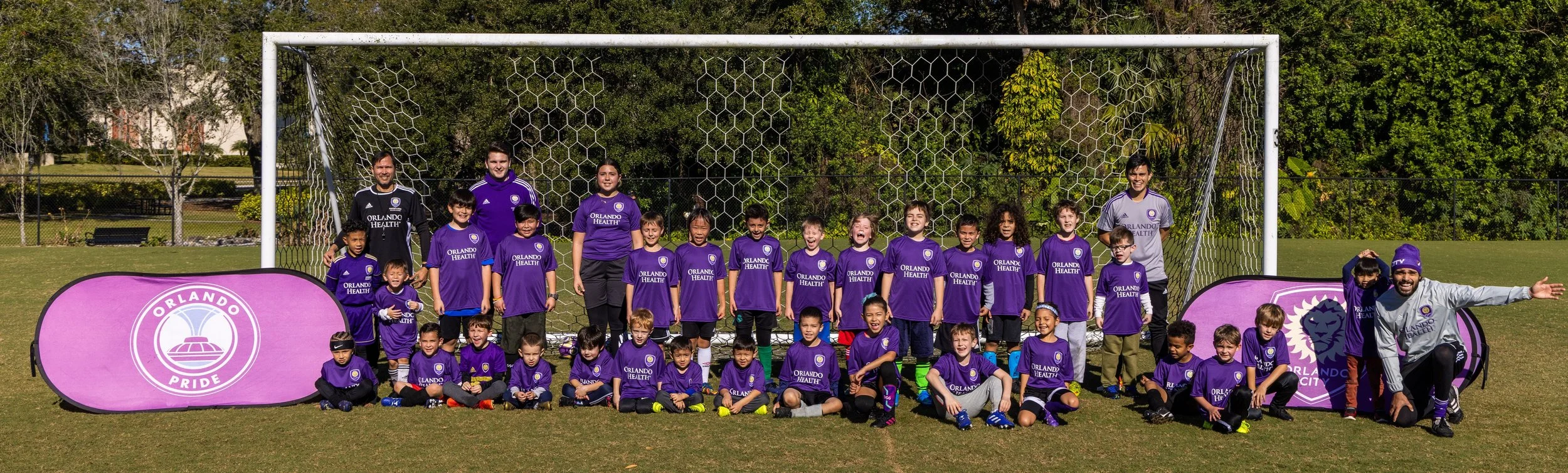 Group of children and adults wearing purple Orlando Pride soccer jerseys posing on a soccer field with banners in front of a goal.