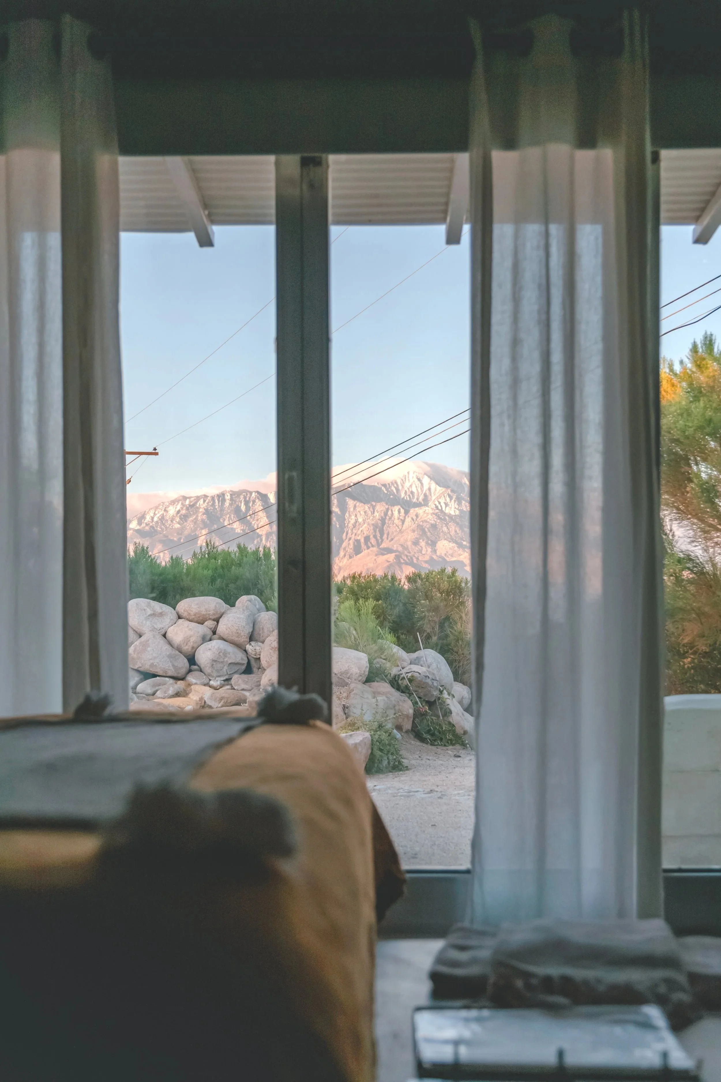 View from inside a room looking out through a window with sheer curtains at a rocky landscape and mountain range with some snow on the peaks, during daylight.