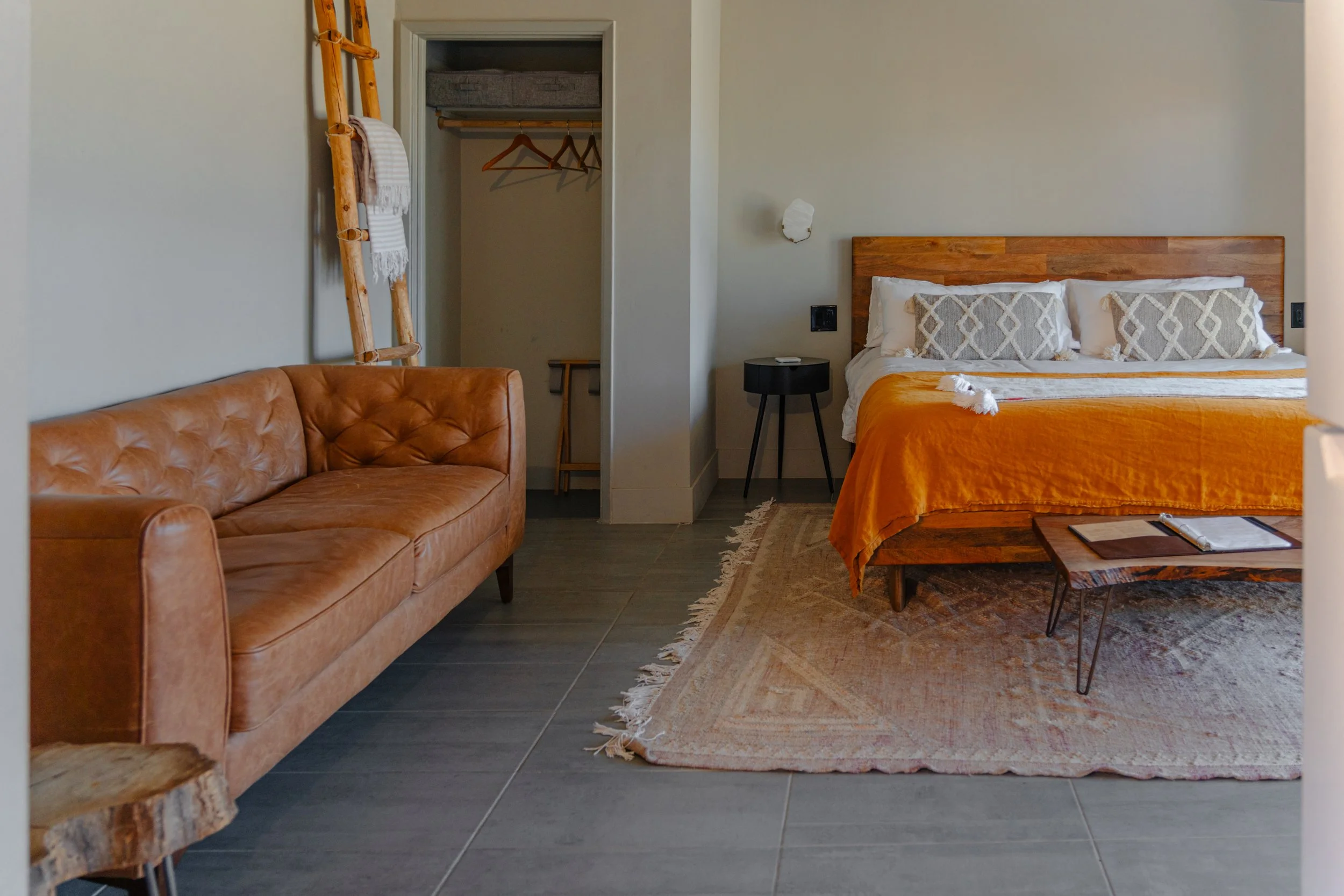 Hotel room with a leather sofa, a bed with an orange blanket, a small black side table, and an open closet with hanging clothes and a ladder-like decor piece.