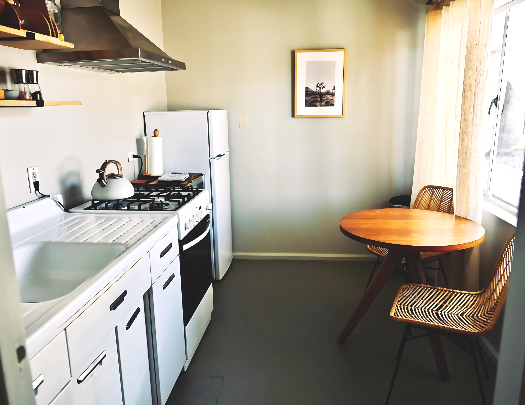 Small kitchen with white cabinets, a white sink, a stovetop with a kettle, and a white refrigerator. There is a round wooden dining table with two woven chairs near a large window with cream curtains. A framed black-and-white picture hangs on the green wall.
