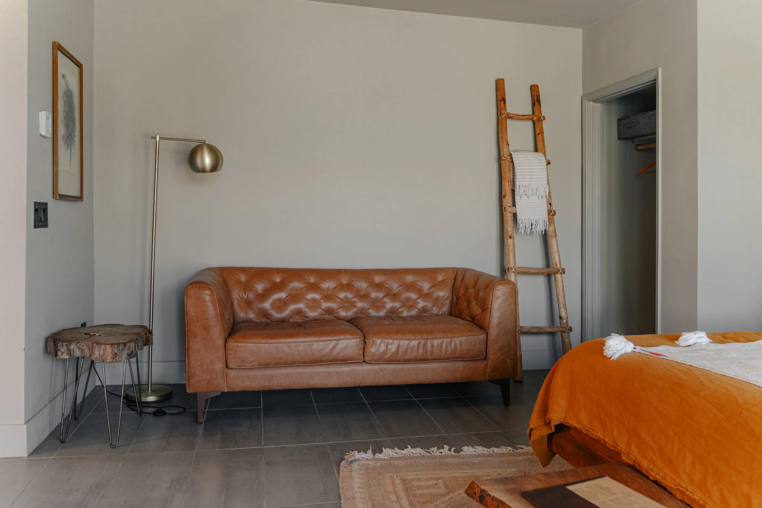 A cozy bedroom corner with a leather sofa, a standing floor lamp, a tree stump side table, a wooden ladder leaning against the wall with a towel, an open closet with clothes, and part of a bed with an orange bedspread.