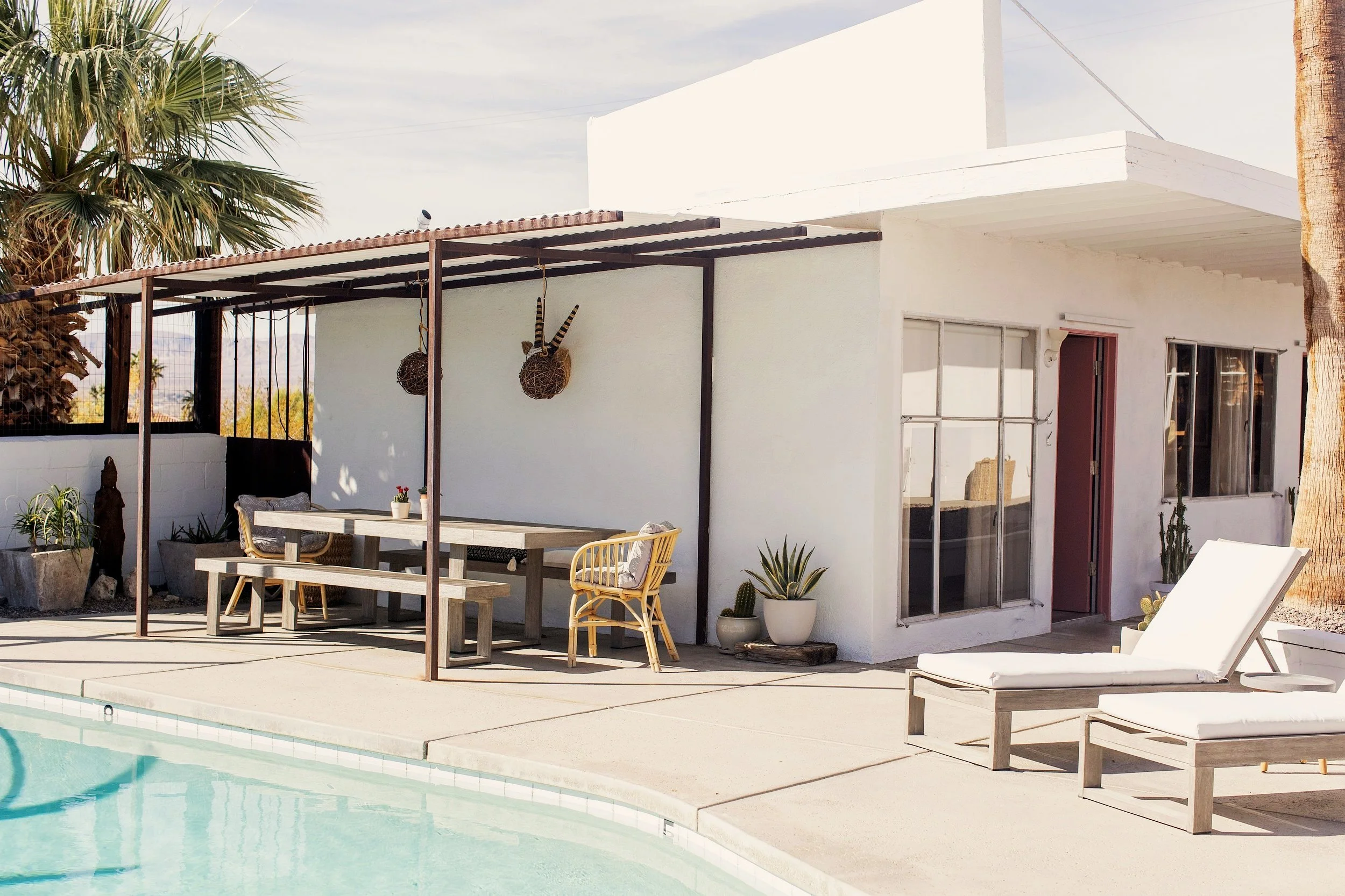 A backyard pool area with white lounge chairs, potted plants, and an outdoor dining table with chairs. The house has large windows and a door, with a shaded patio area overhead, decorated with hanging baskets and a palm tree in the background.