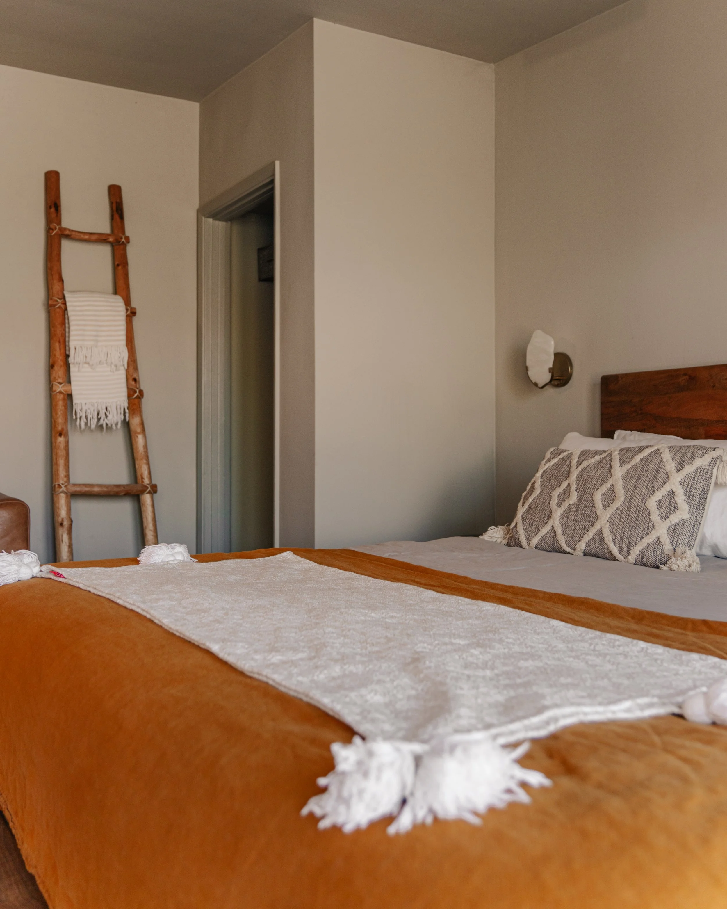A neatly made bed with an orange bedspread and white and beige decorative pillows, a rustic wooden ladder with a white blanket, and a wall-mounted light fixture in a cozy bedroom.