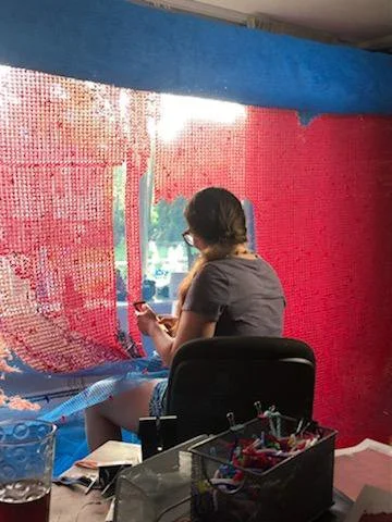 A woman sitting on a chair in front of a multicolored curtain made of small fabric pieces, with a view outside through the window, holding a phone in her hand, with art supplies and a glass of water nearby.