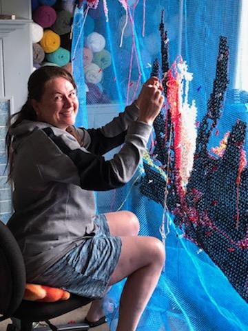 A woman creating a diamond painting artwork on a blue mesh canvas, with colorful beads and thread, sitting on a black chair in a room with yarn storage in the background.