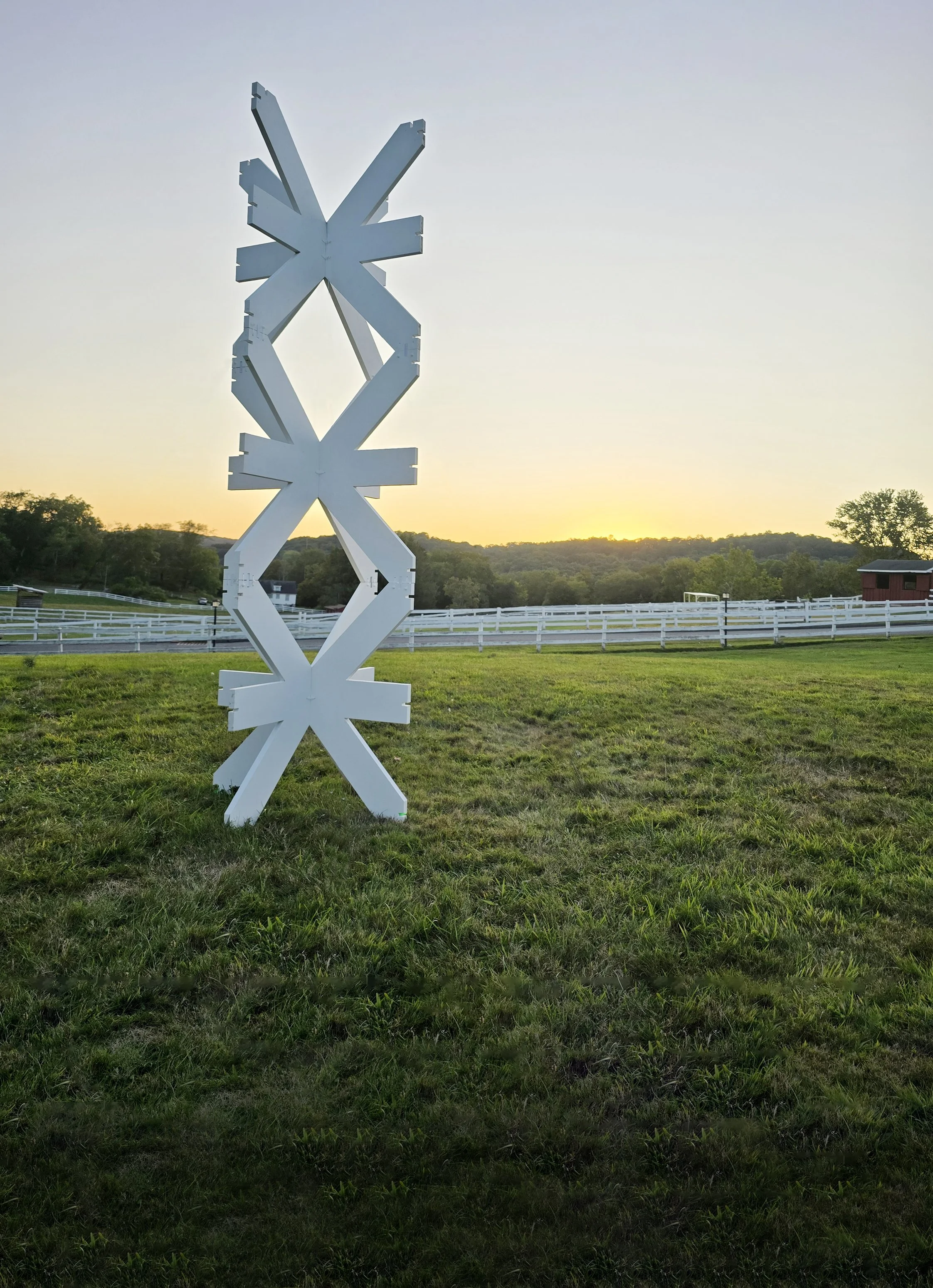 A large white modern sculpture made of geometric shapes stands in a grassy field at sunset. A white fence and trees are visible in the background.