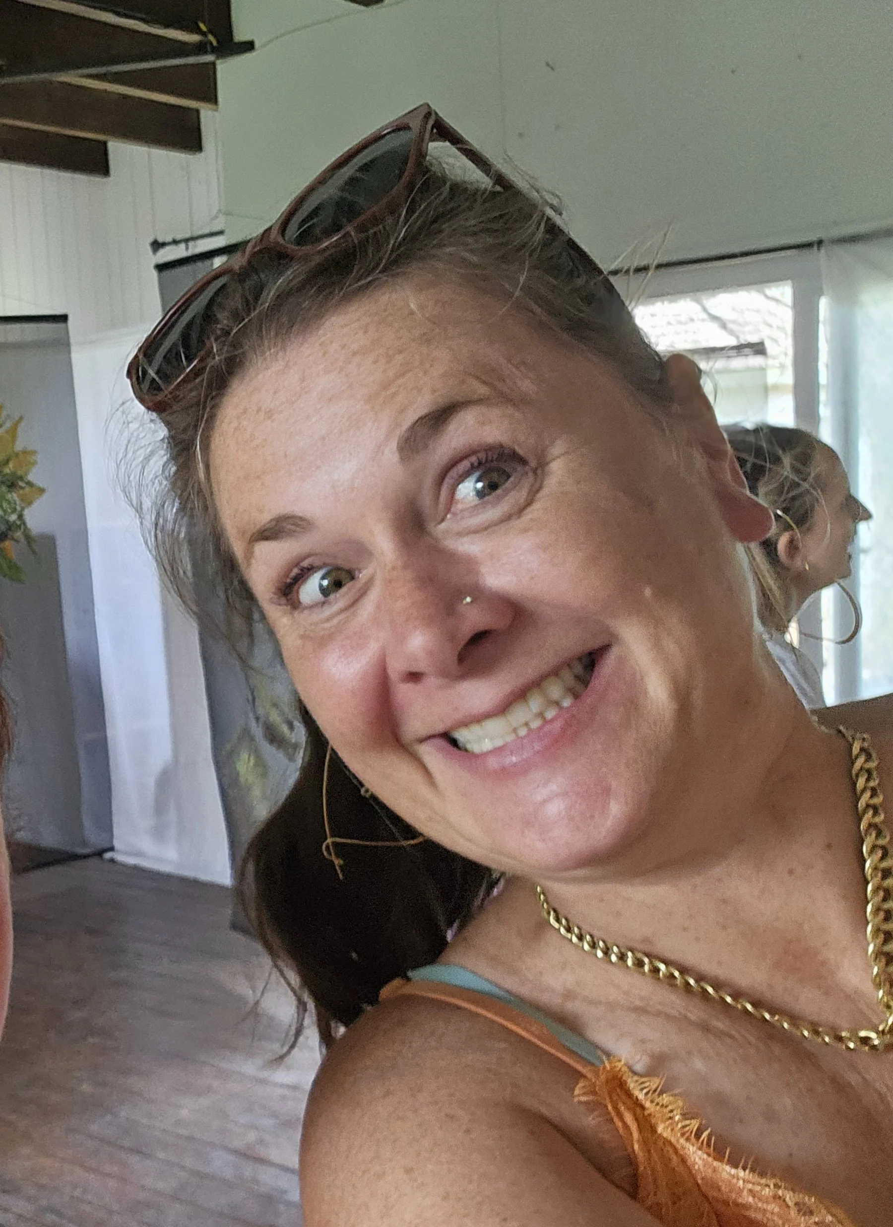 Close-up of a woman smiling with sunglasses on her head and a gold necklace, indoors with another person blurred in the background.