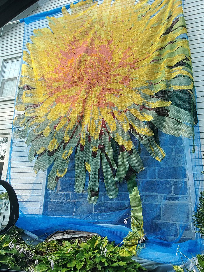 A large sunflower mosaic artwork being attached to the exterior wall of a house, covered with protective blue netting.