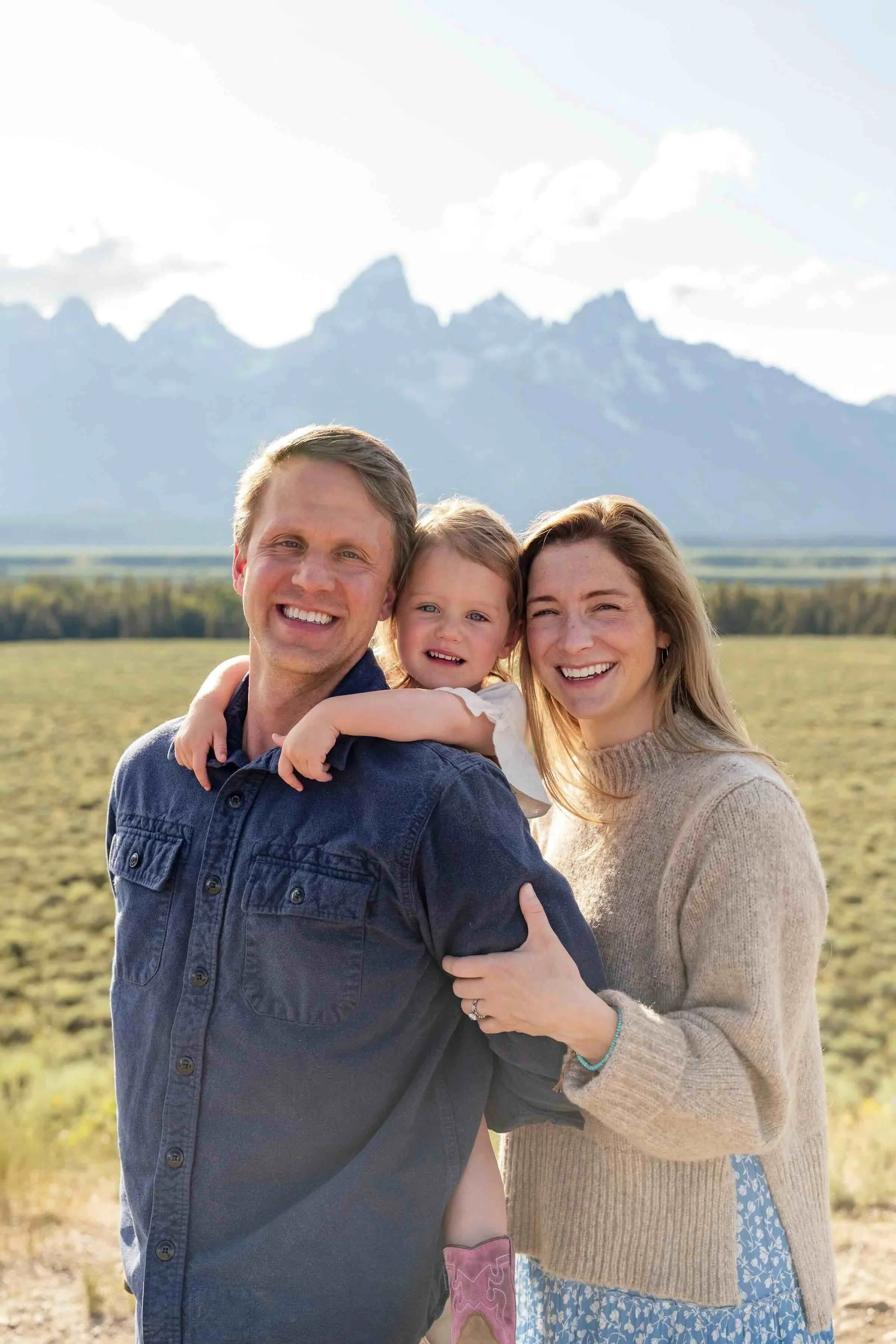 A smiling family of three, consisting of a father, mother, and young daughter, standing outdoors in a field with mountains in the background during daytime.