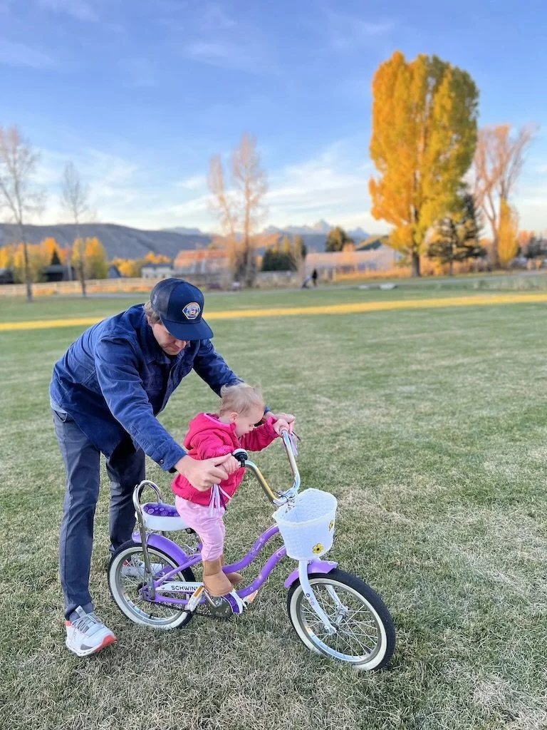 greg hahnel teaching a young girl how to ride a purple and white bicycle with a small white basket in a park during sunset, with yellow fall trees and mountains in the background.