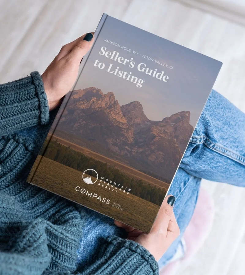 A person holding a real estate guidebook titled 'Seller's Guide to Listing' with mountain scenery on the cover, wearing a blue knit sweater and jeans.