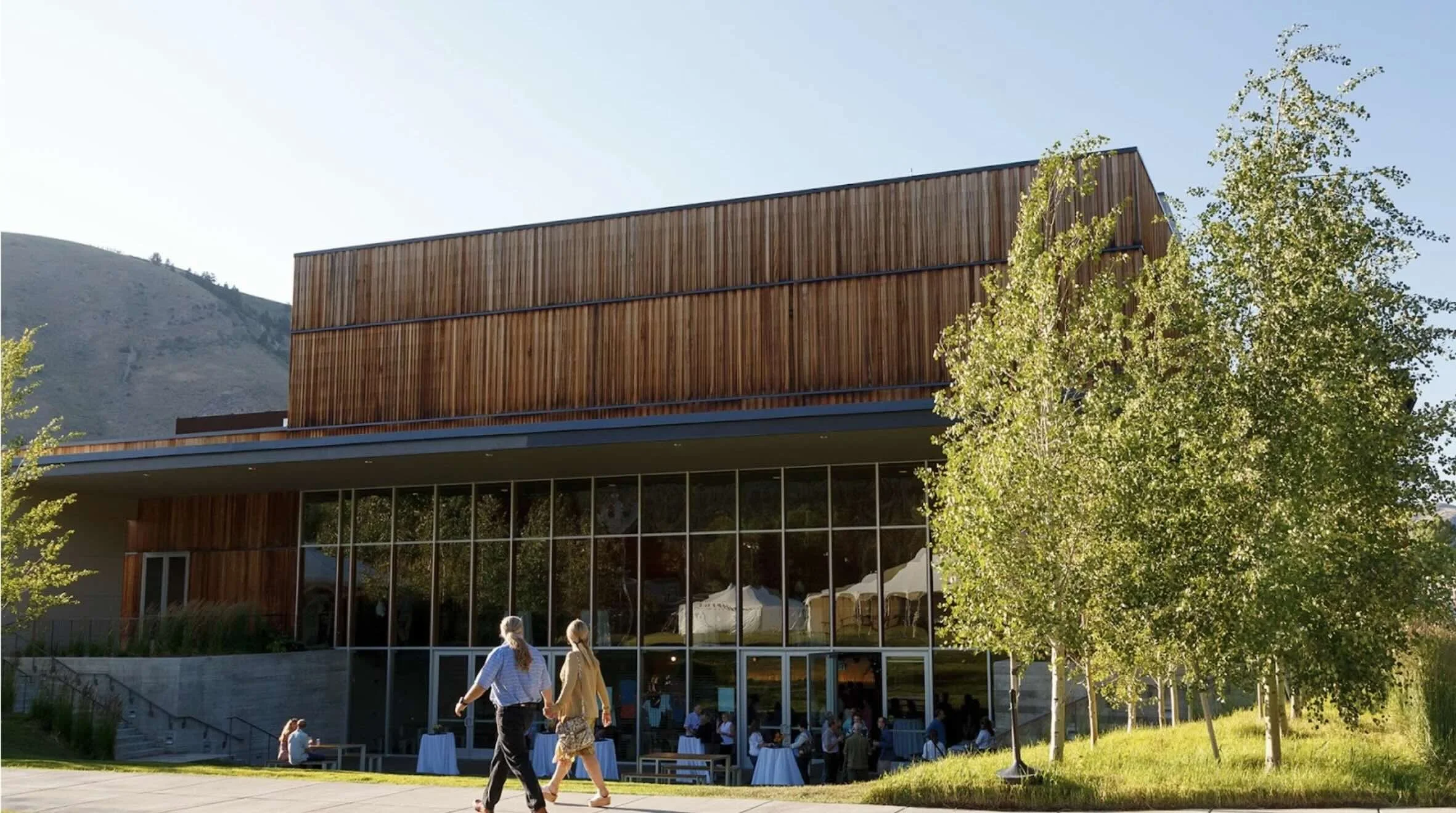 A man and a woman holding hands walking in front of the Center for the Arts on a summer day.