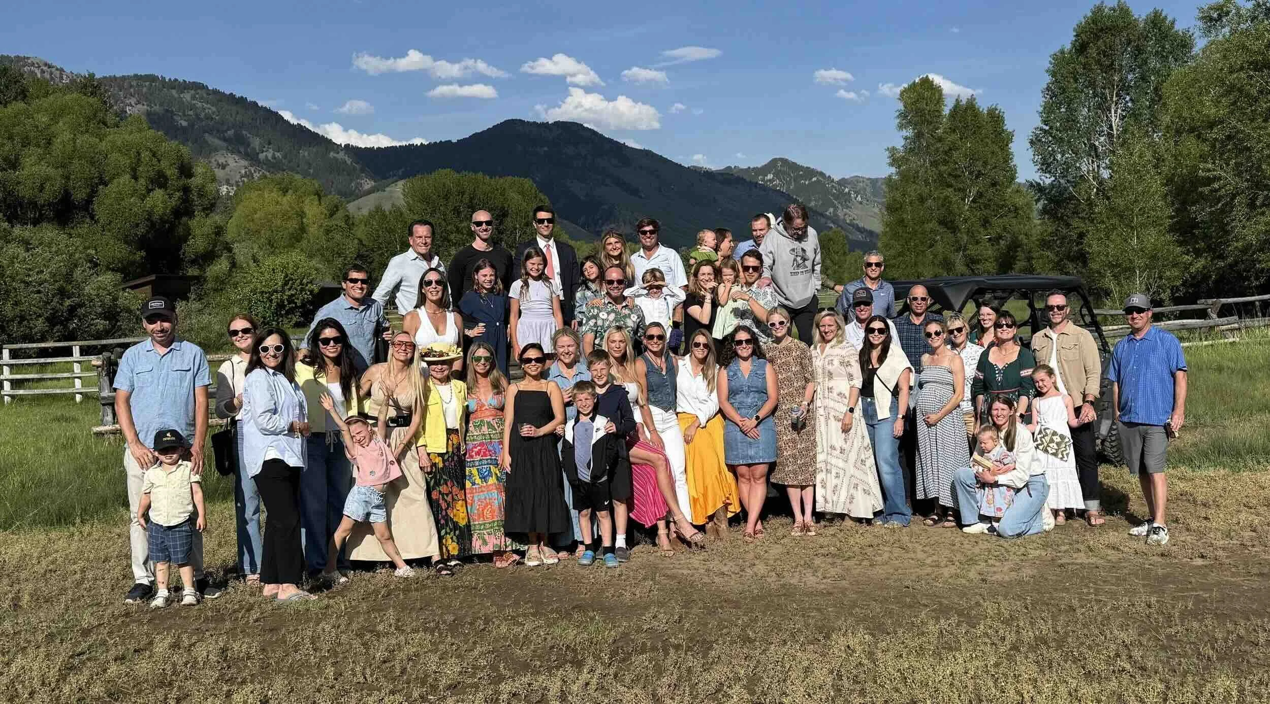 A large group of people gathered outdoors on a sunny day with mountains and trees in the background, posing for a group photo.