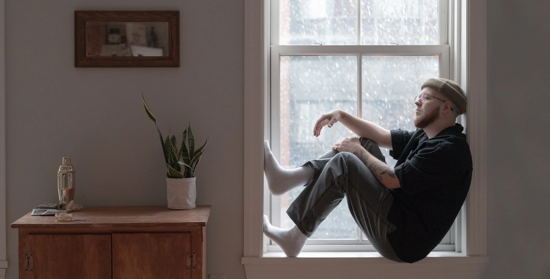 Person sitting on a windowsill, wearing a beanie and glasses, looking outside. Next to the window is a wooden cabinet with a potted plant and a metallic bottle on top.