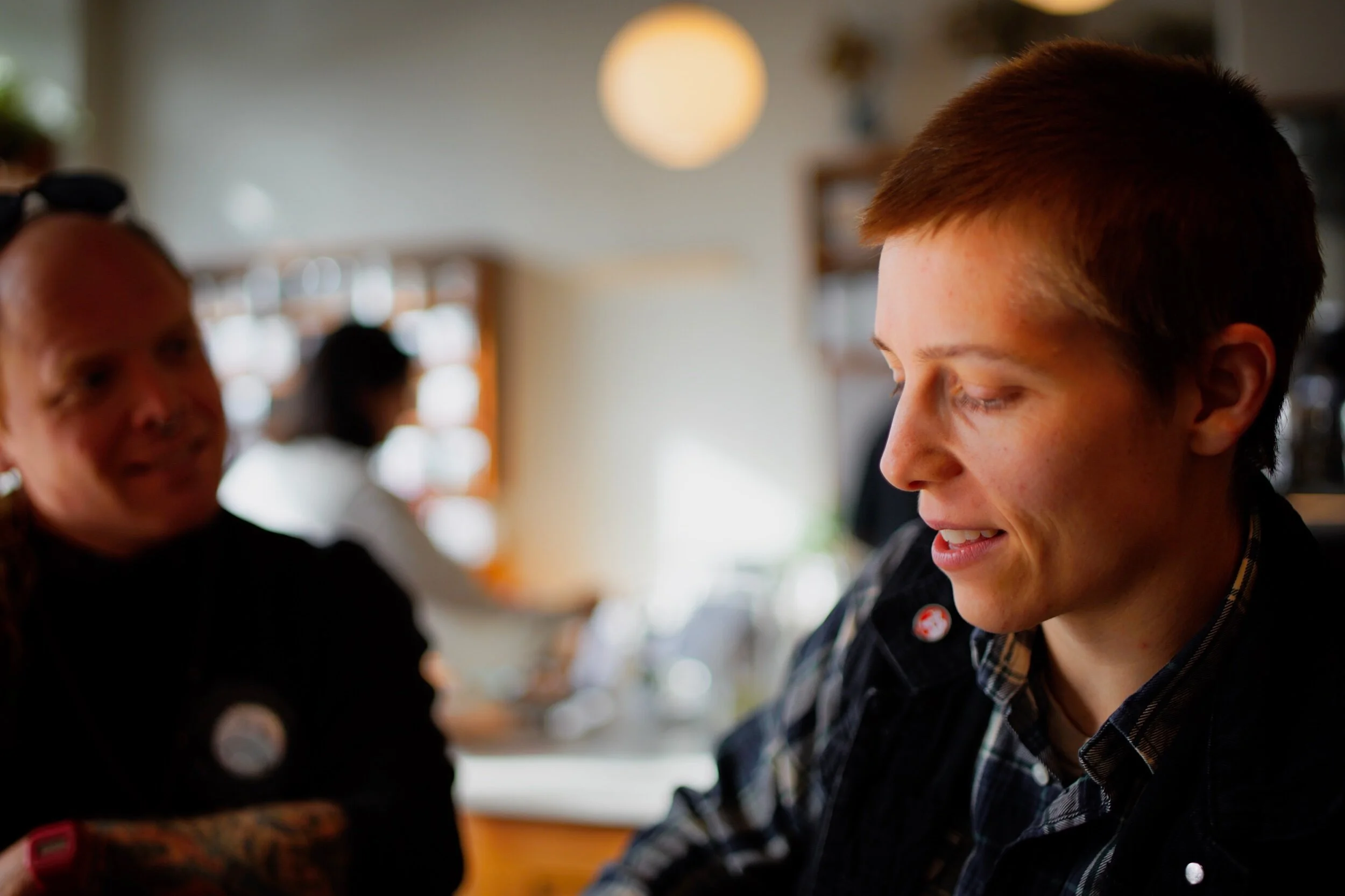 A young person with short brown hair smiling and looking down, in focus. Another person blurred in the background, in a kitchen or home setting.