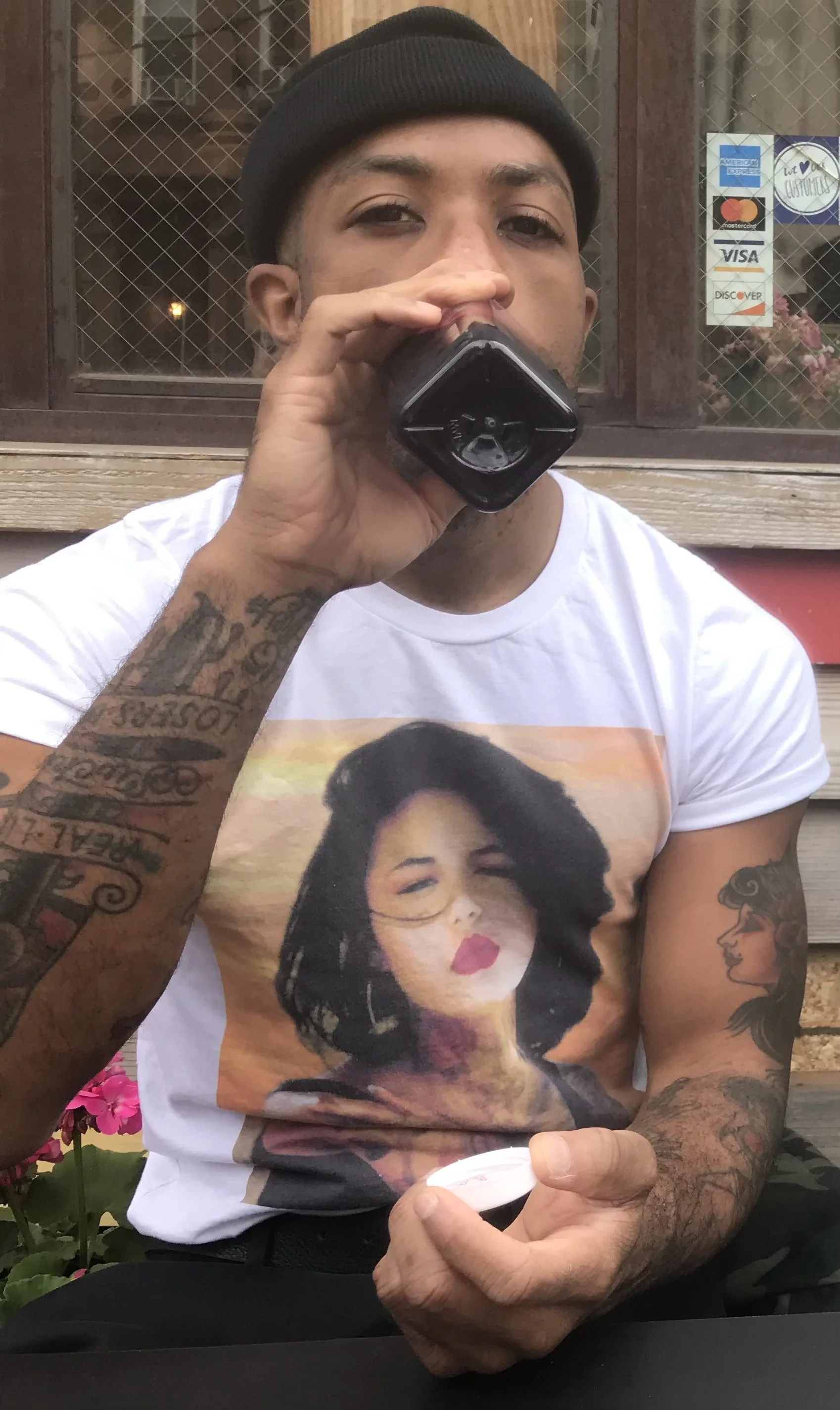 A man wearing a black beanie and a white T-shirt with a woman's face on it, sitting outdoors, drinking from a black bottle, with colorful tattoos on his arms.
