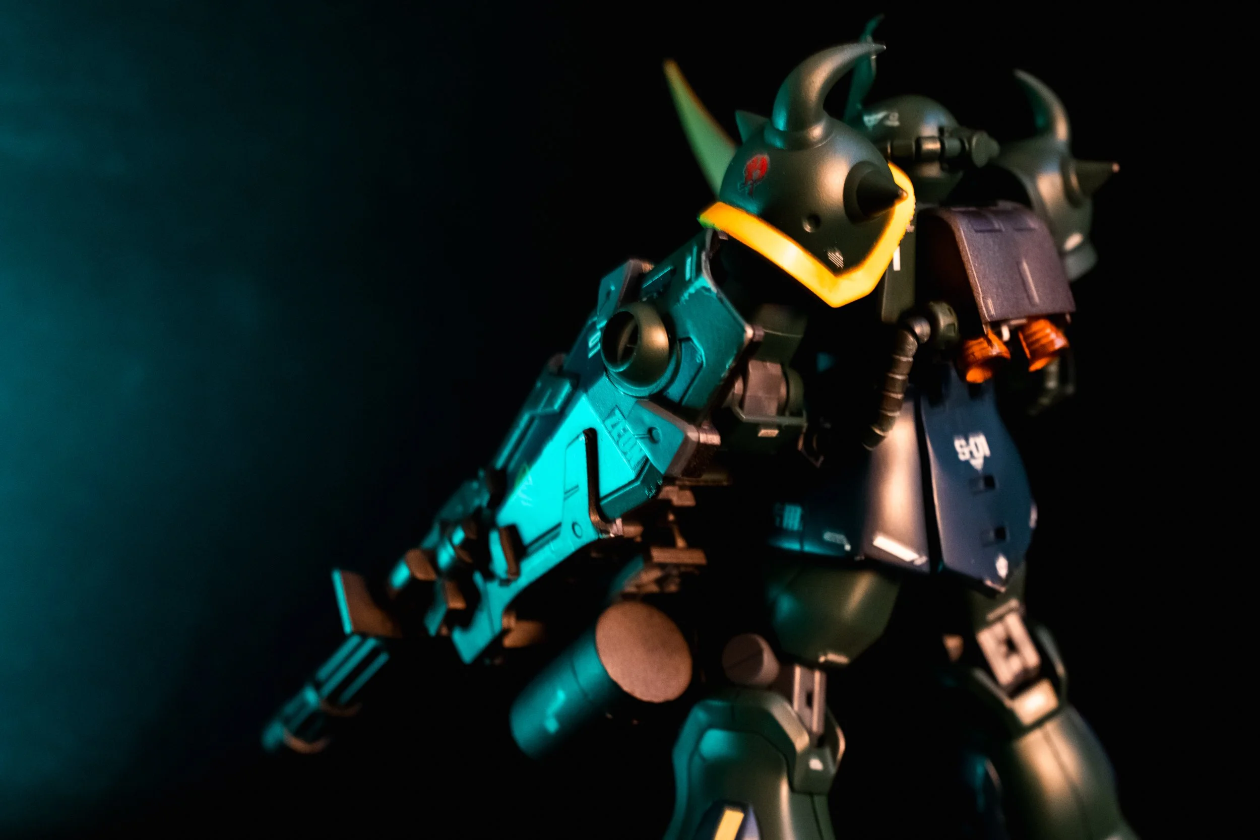 A detailed action figure of a robot or mecha with a helmet featuring a yellow faceplate, large horns, a gun in its right hand, and various armor and mechanical details, set against a dark background with colorful lighting.