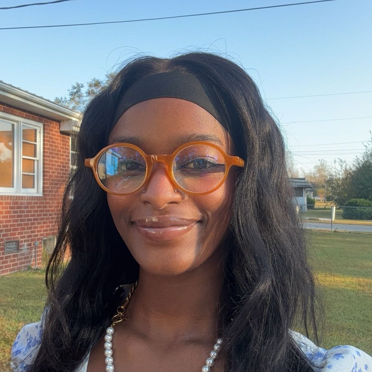 Woman wearing orange glasses, black headband, and a pearl necklace standing outside near a brick house.