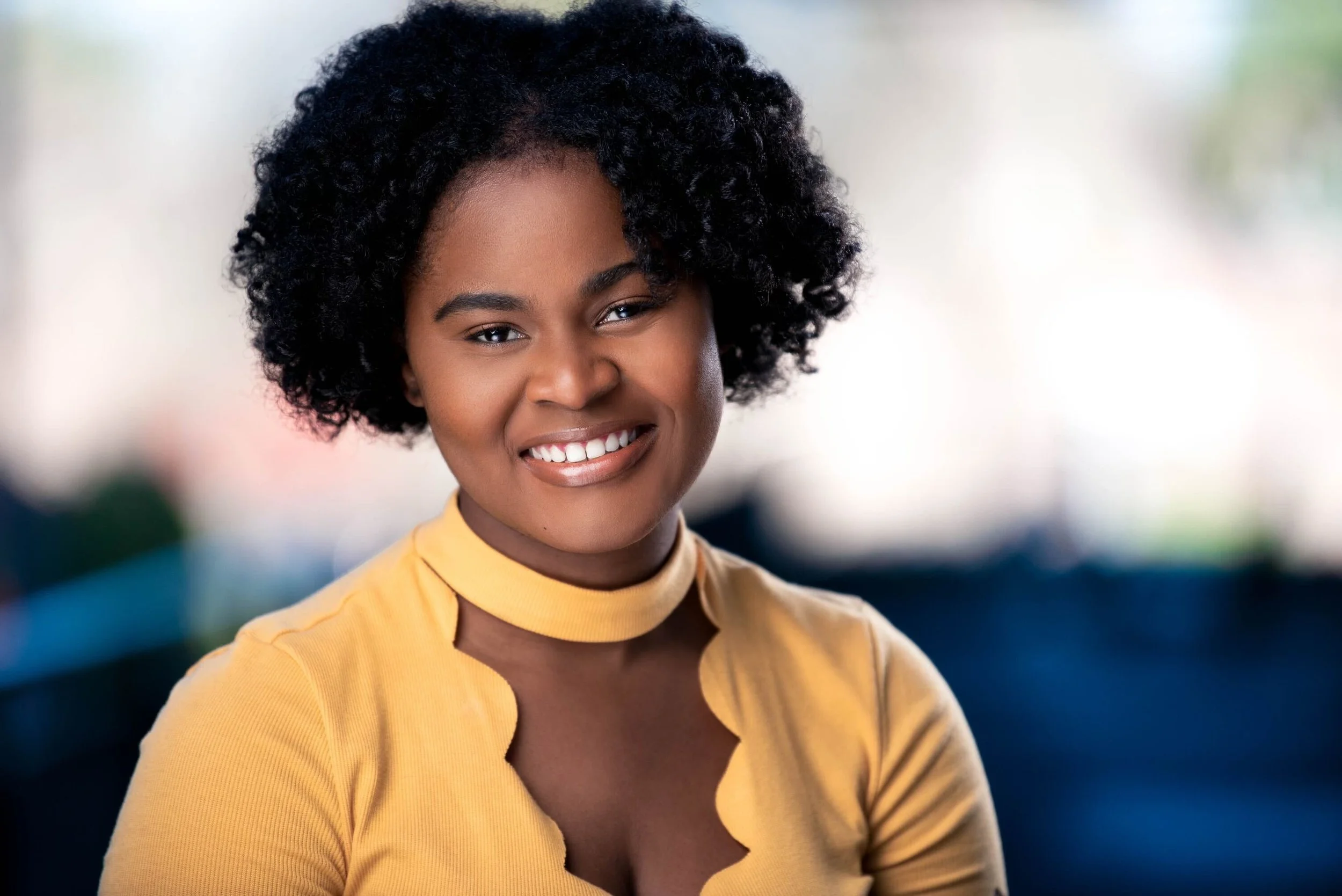 Portrait of person smiling, wearing a yellow top, with natural curly hair.