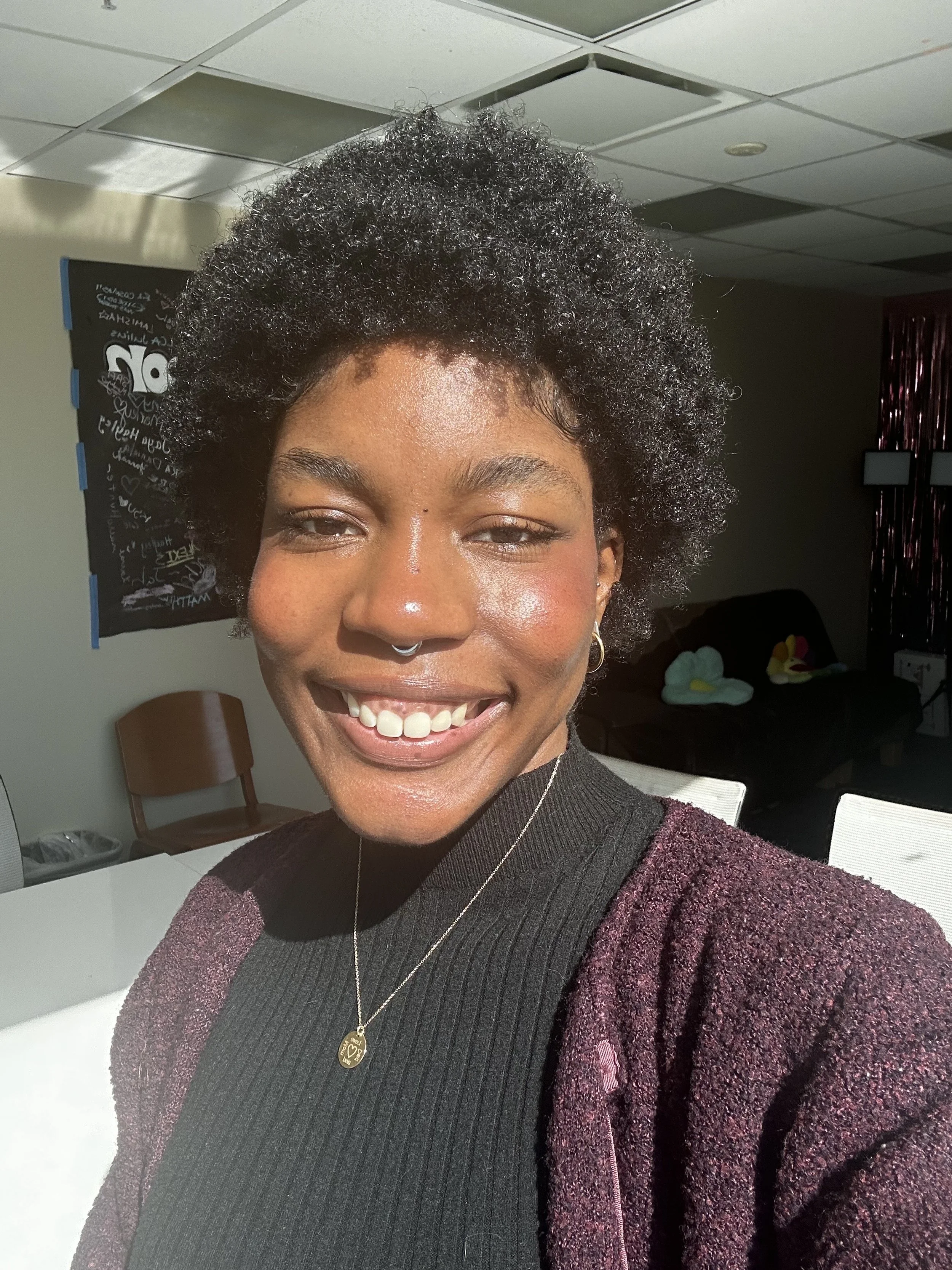 A person smiling brightly in an indoor setting, wearing a black top, purple cardigan, and a necklace. The background includes a chair and wall decor, with sunlight illuminating their face.