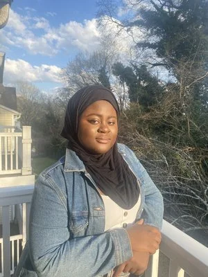 Person wearing a hijab and denim jacket standing on a balcony with a green and cloudy sky backdrop.
