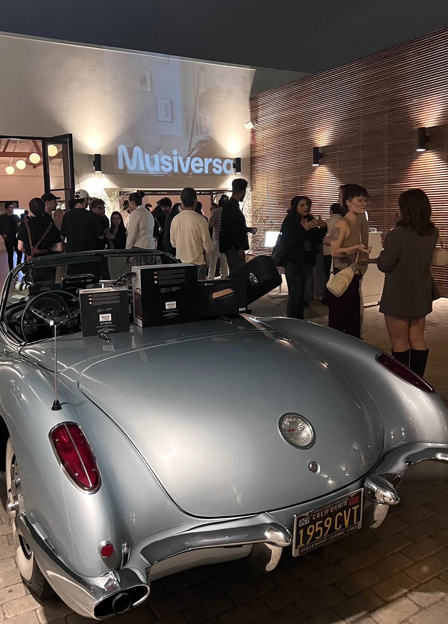 A silver classic car with a California license plate, parked in front of a crowded building entrance with people socializing, some holding drinks, and a sign that reads 'Musiversa' on the wall.