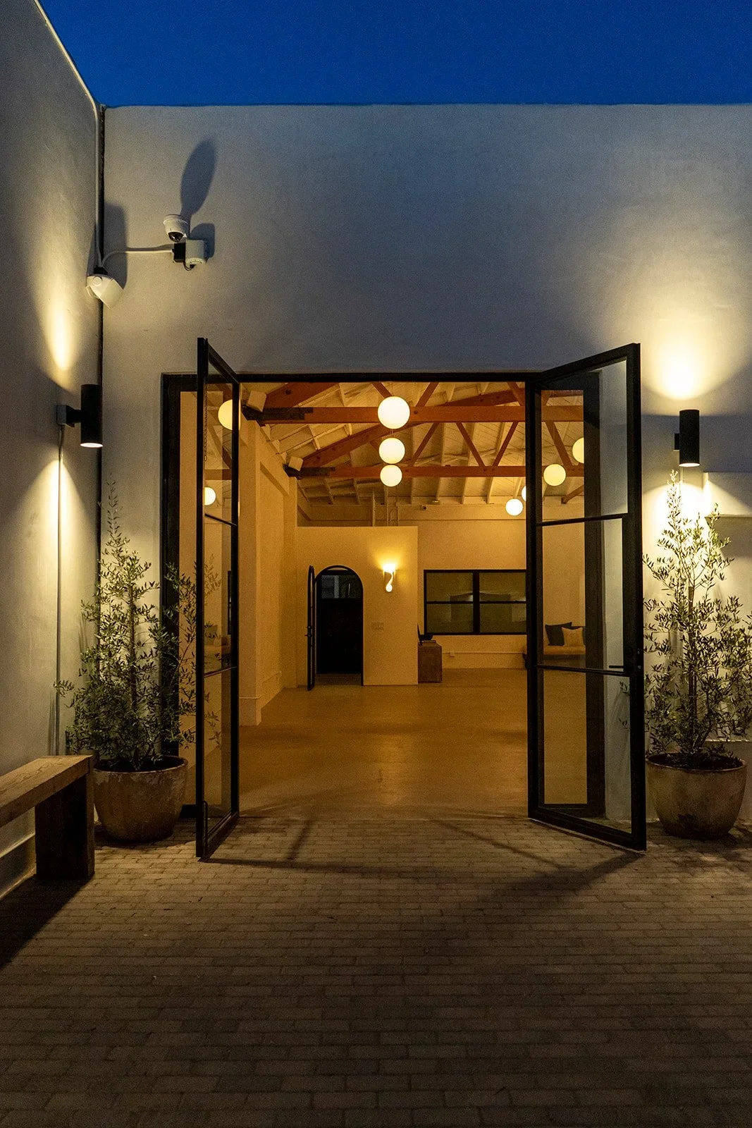 Lacuna indoor outdoor event space entry through open french doors at dusk with bow truss ceiling and globe lighting West Adams Los Angeles
