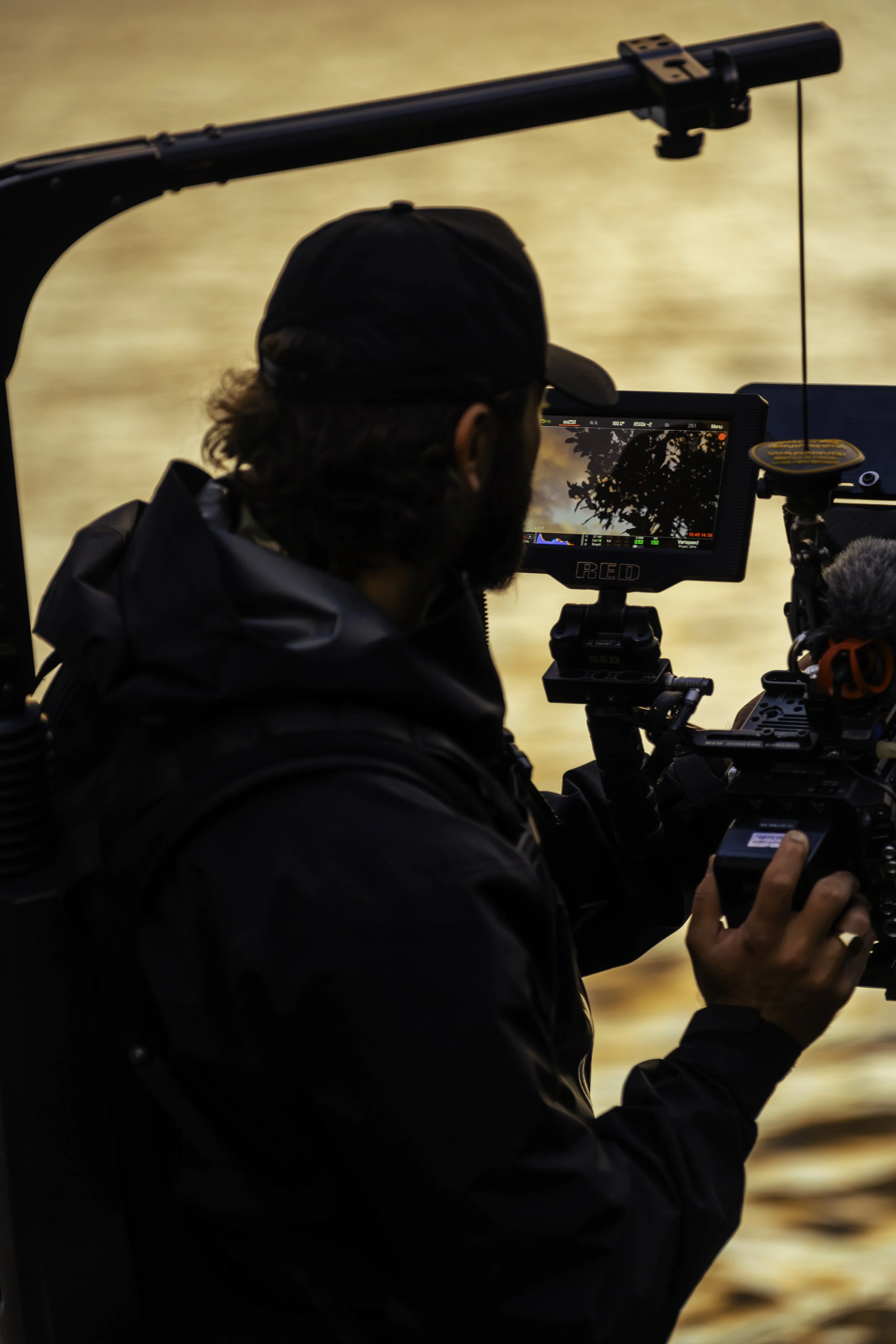 A filmmaker is filming scenery with a professional camera, wearing a black cap and jacket, during sunset or sunrise.