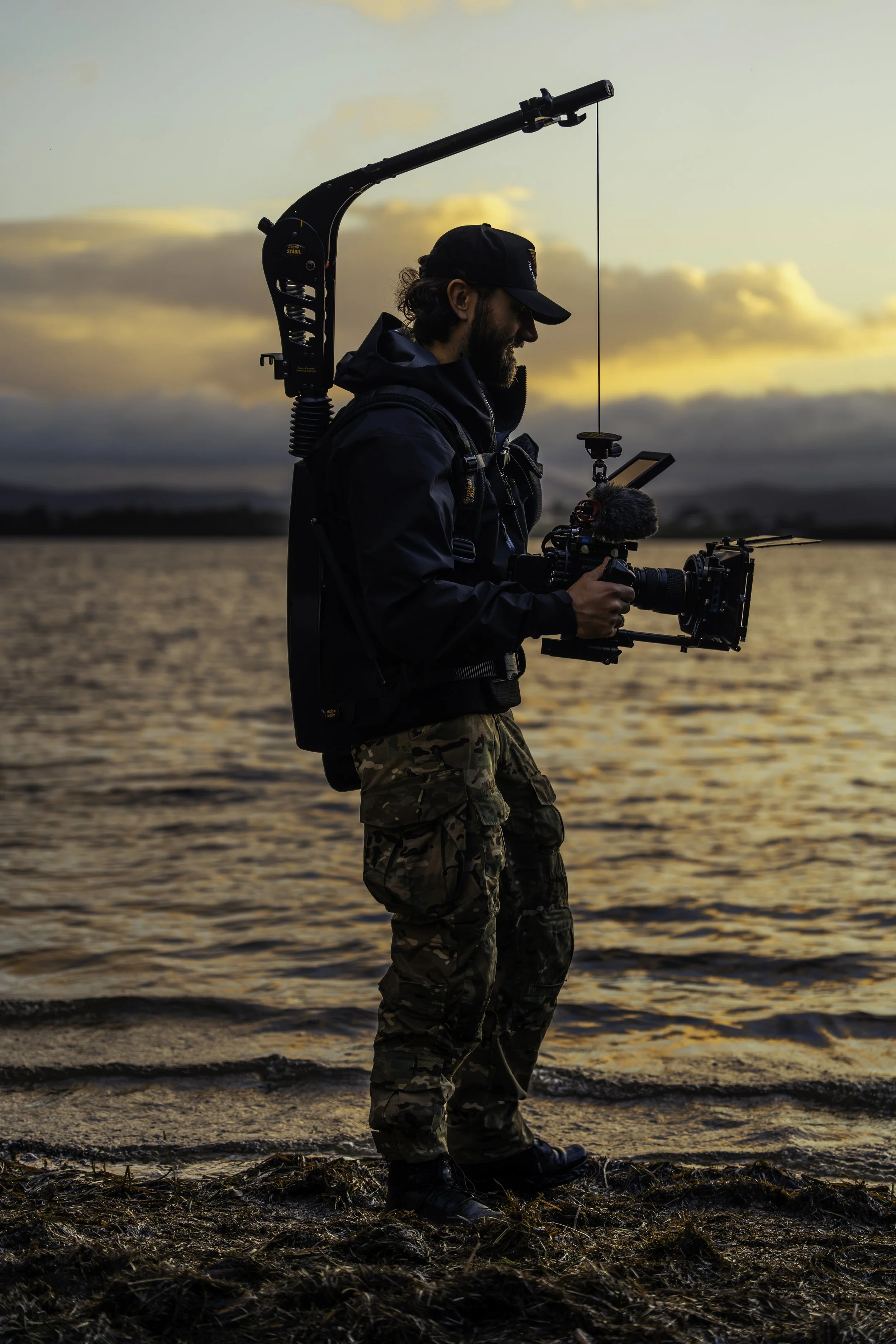 A man with a beard and curly hair standing on the beach during sunset, wearing camouflage pants, a dark jacket, and a hat, while operating professional video equipment with a stabilizer, microphone, and monitor.