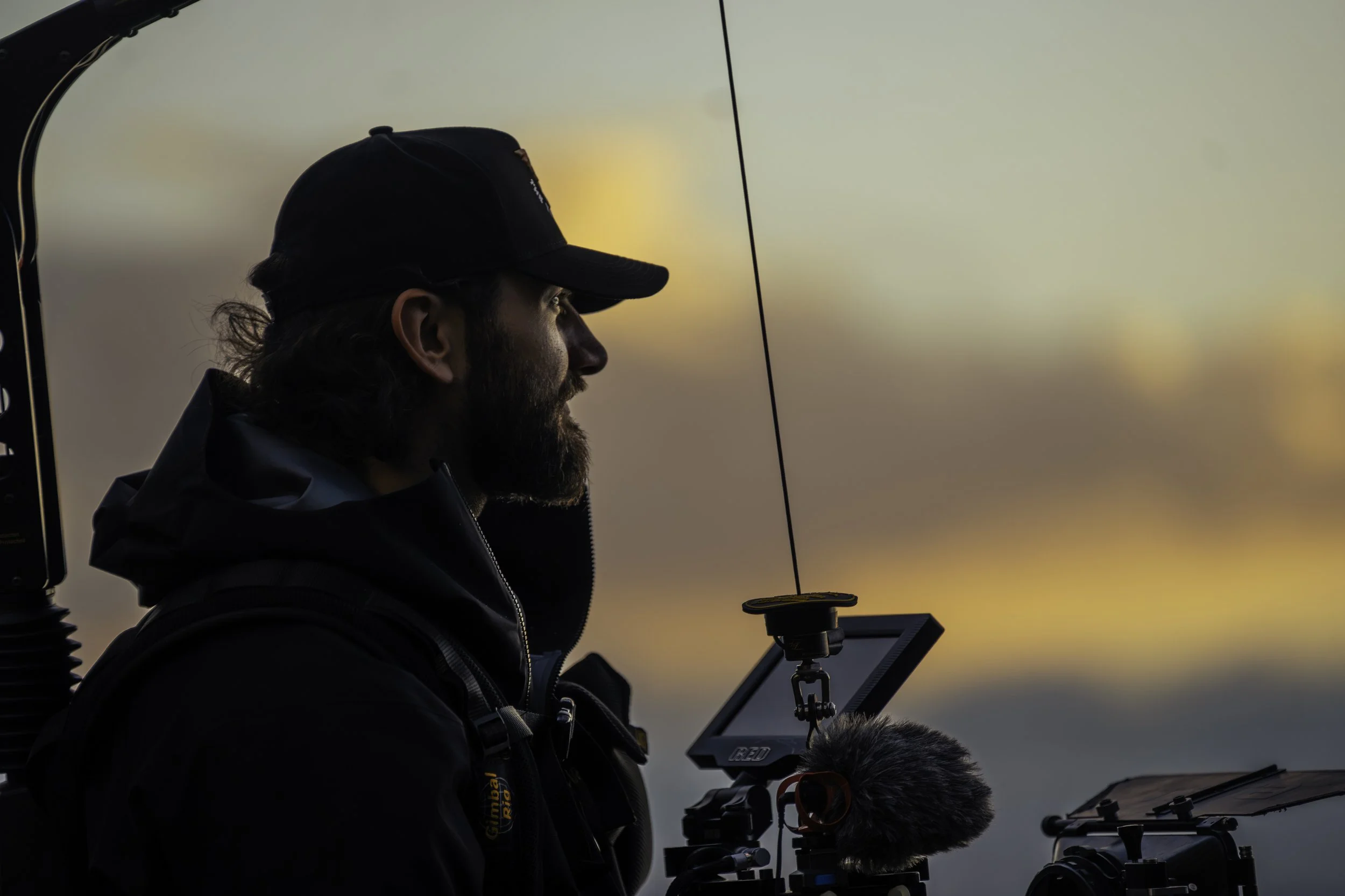 A man with long hair and a beard wearing a black cap and black jacket, operating a drone with a remote control device, during sunset or sunrise with a blurry sky in the background.