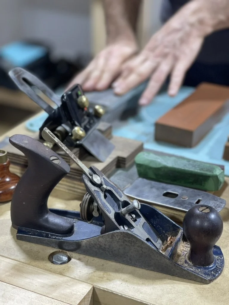 Hand plane and sharpening stones on the bench at Moonlite Studios, with hands working a plane iron on a waterstone