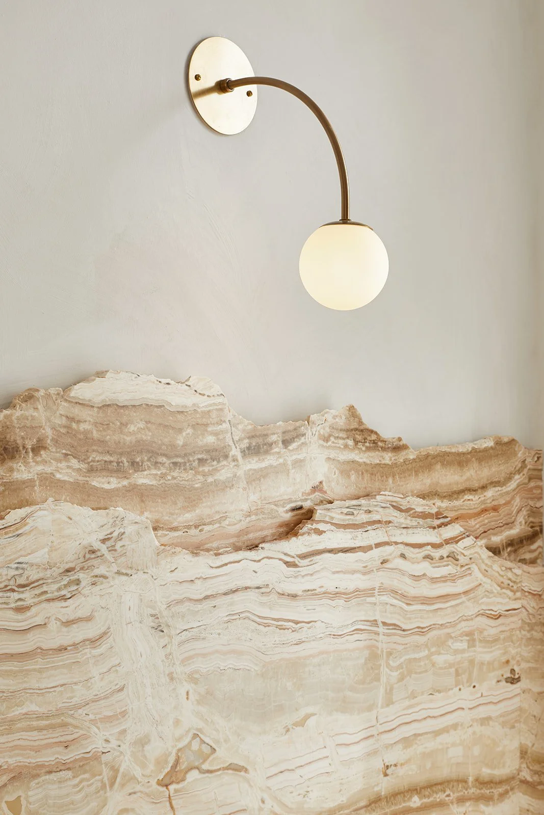 A modern wall-mounted light fixture with a white glass globe shade and an arched brass arm, positioned above a textured beige and brown marble surface.