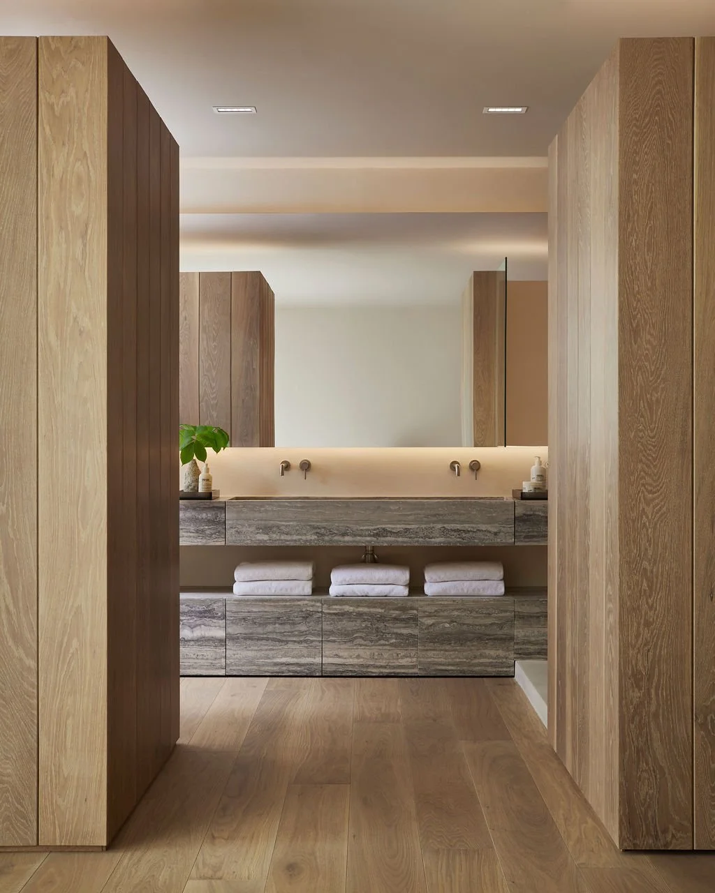 A symmetrical view of a serene primary bathroom featuring a custom floating travertine stone double vanity, framed by floor-to-ceiling ribbed oak millwork and soft backlit mirrors.