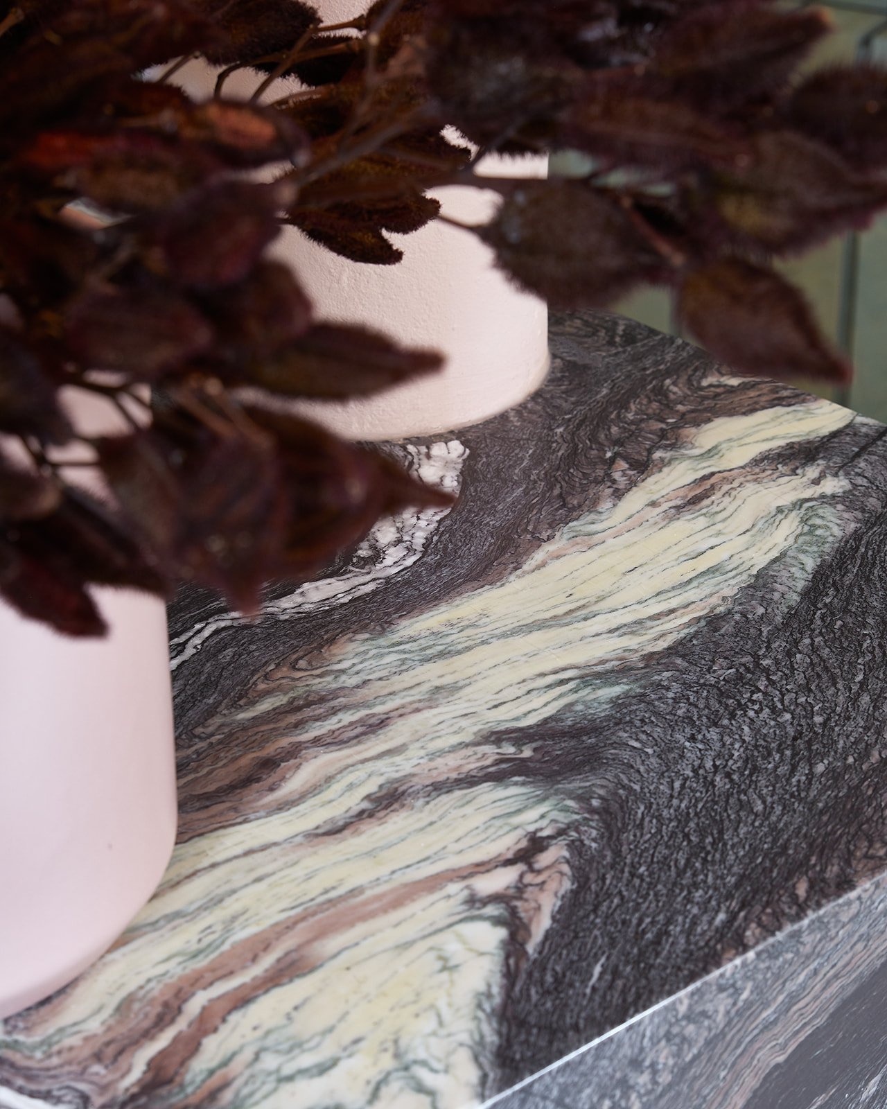 Close-up of dark purple flowers in a pink pot on a marble surface with a white object and a blurred background.